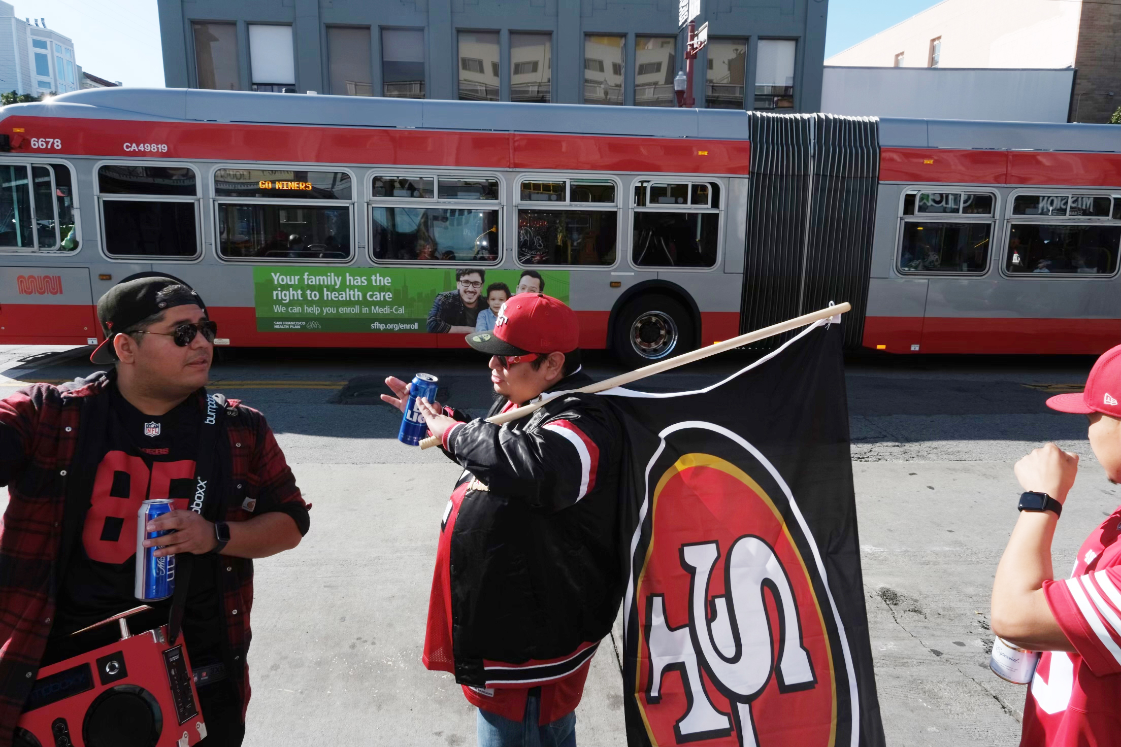 Three 49ers fans in jerseys on a street, one holding a flag, with a red bus behind them.