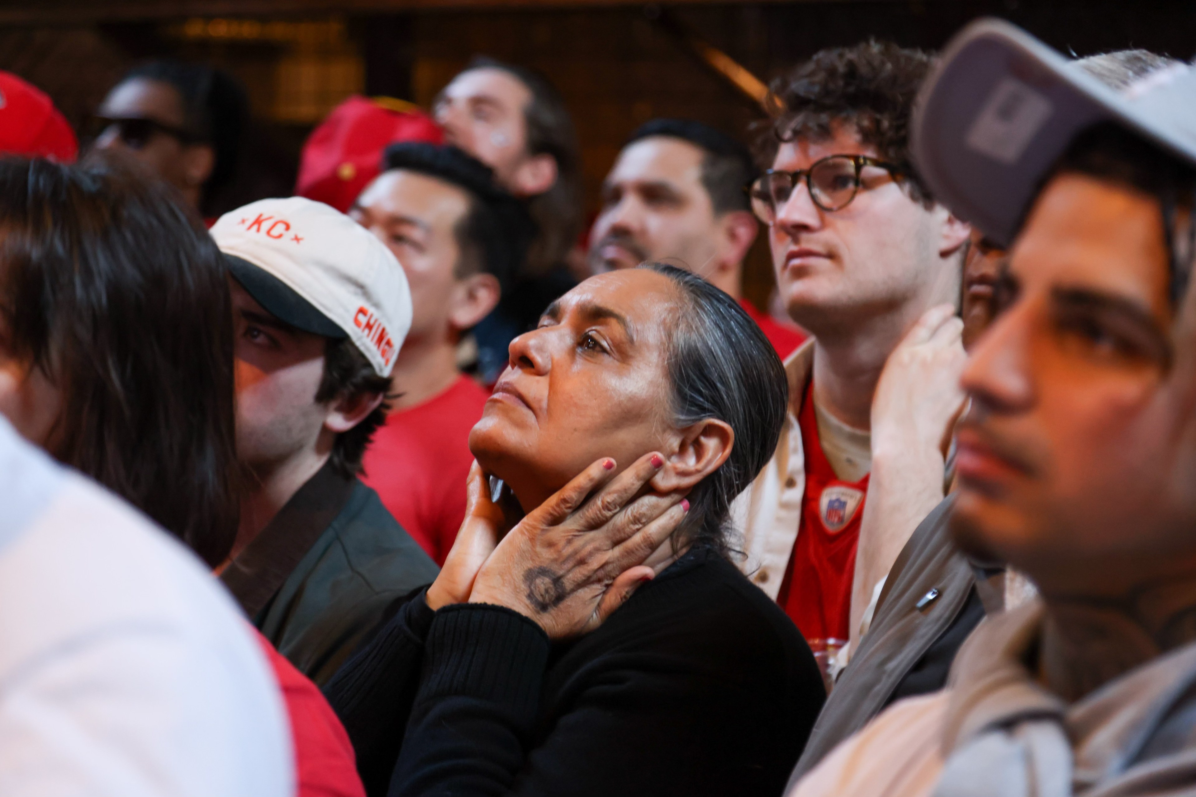 A crowd of people, intently looking upwards, with some wearing red attire and caps. A woman stands out with her hands clasped at her neck.