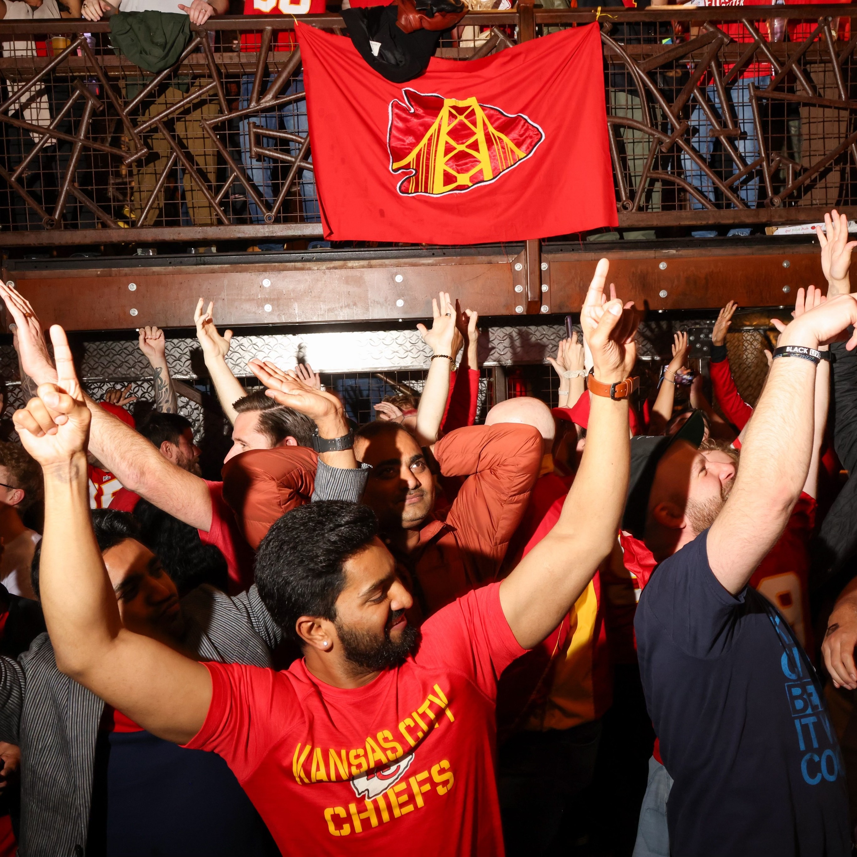 A crowd of excited people are cheering with their hands up, many wearing Kansas City Chiefs shirts, surrounded by red banners and a lively atmosphere.