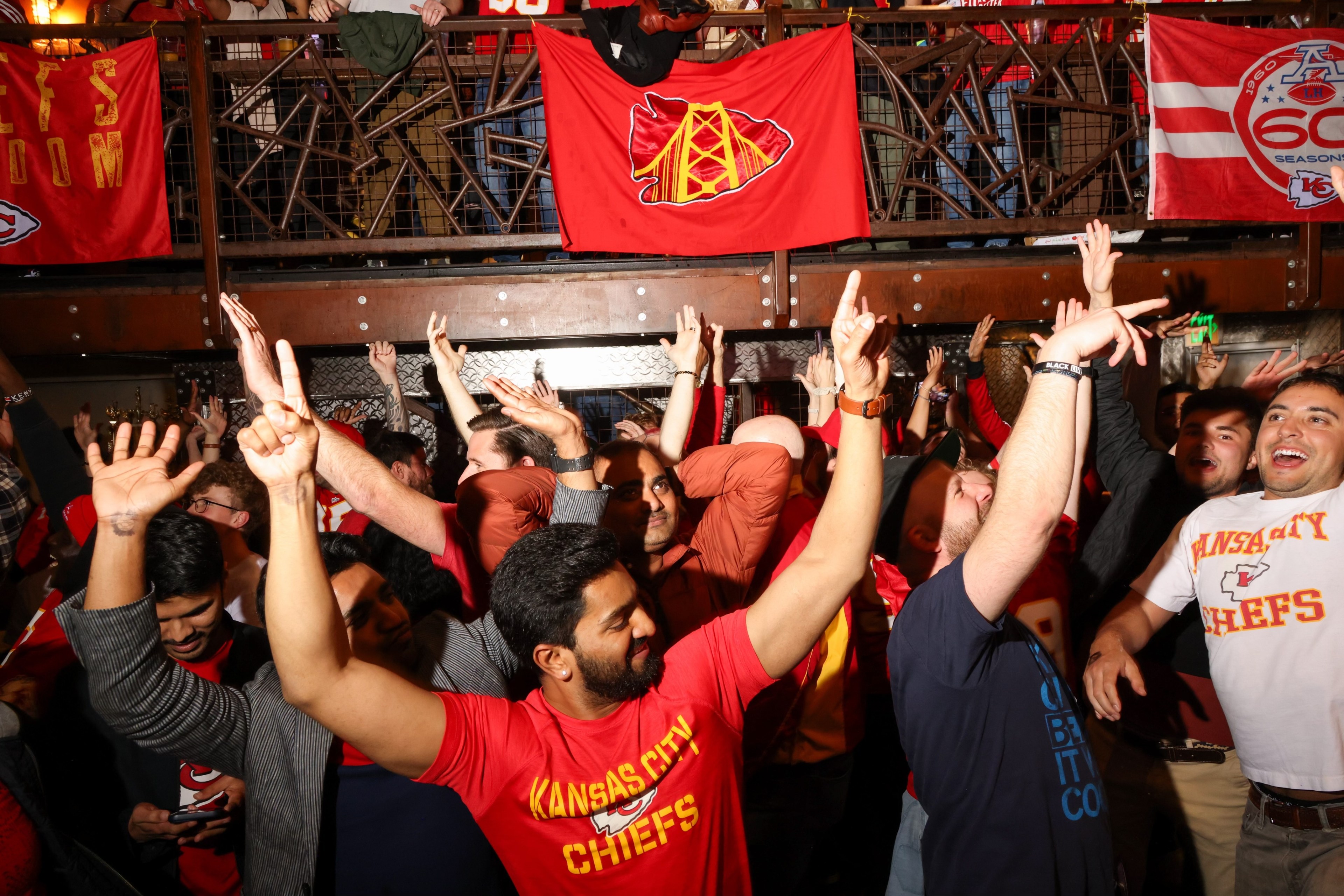 A crowd of excited people are cheering with their hands up, many wearing Kansas City Chiefs shirts, surrounded by red banners and a lively atmosphere.