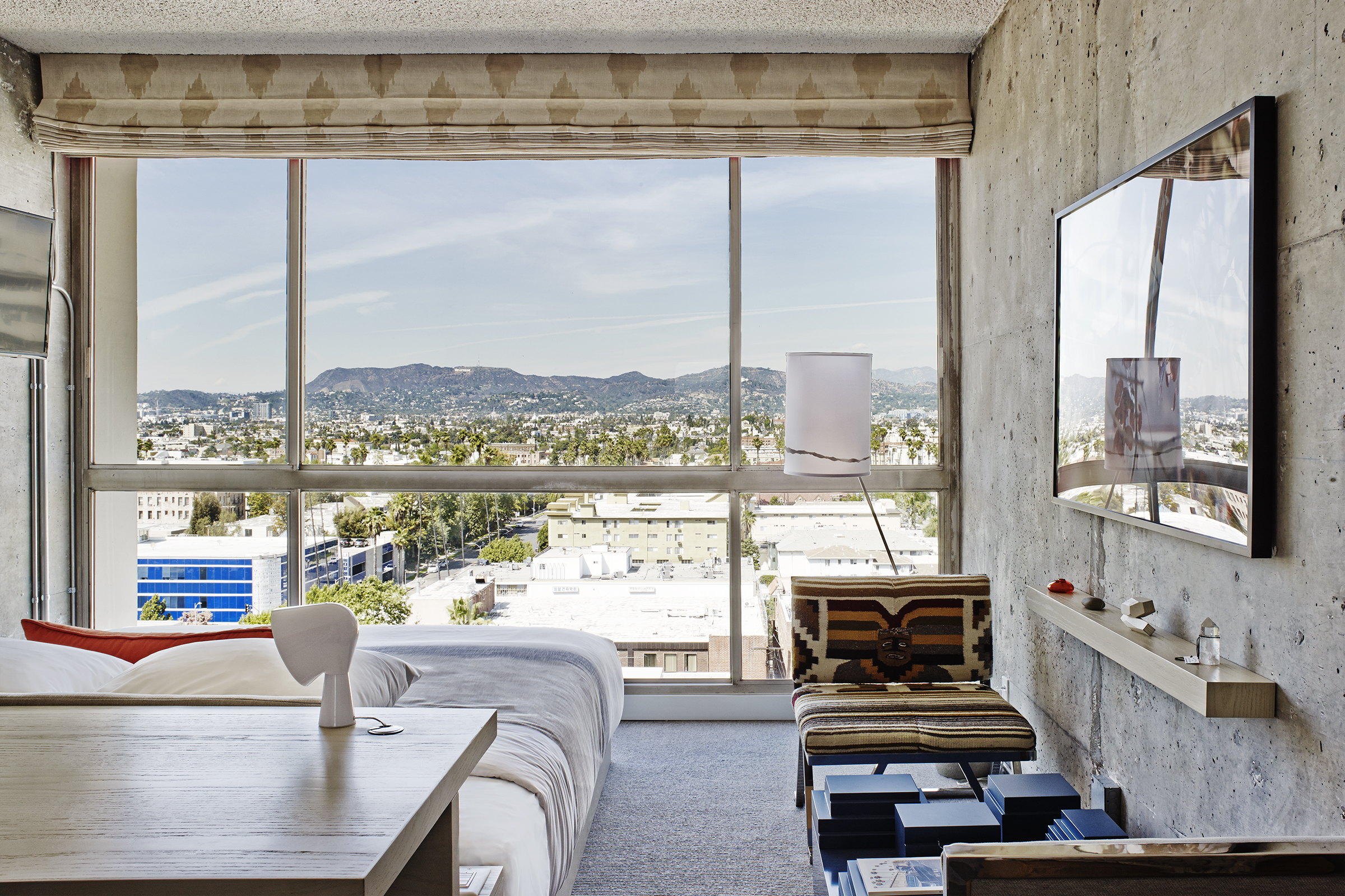 A room with a floor to ceiling window looks out on a cityscape scattered with palm trees and mountains in the distance