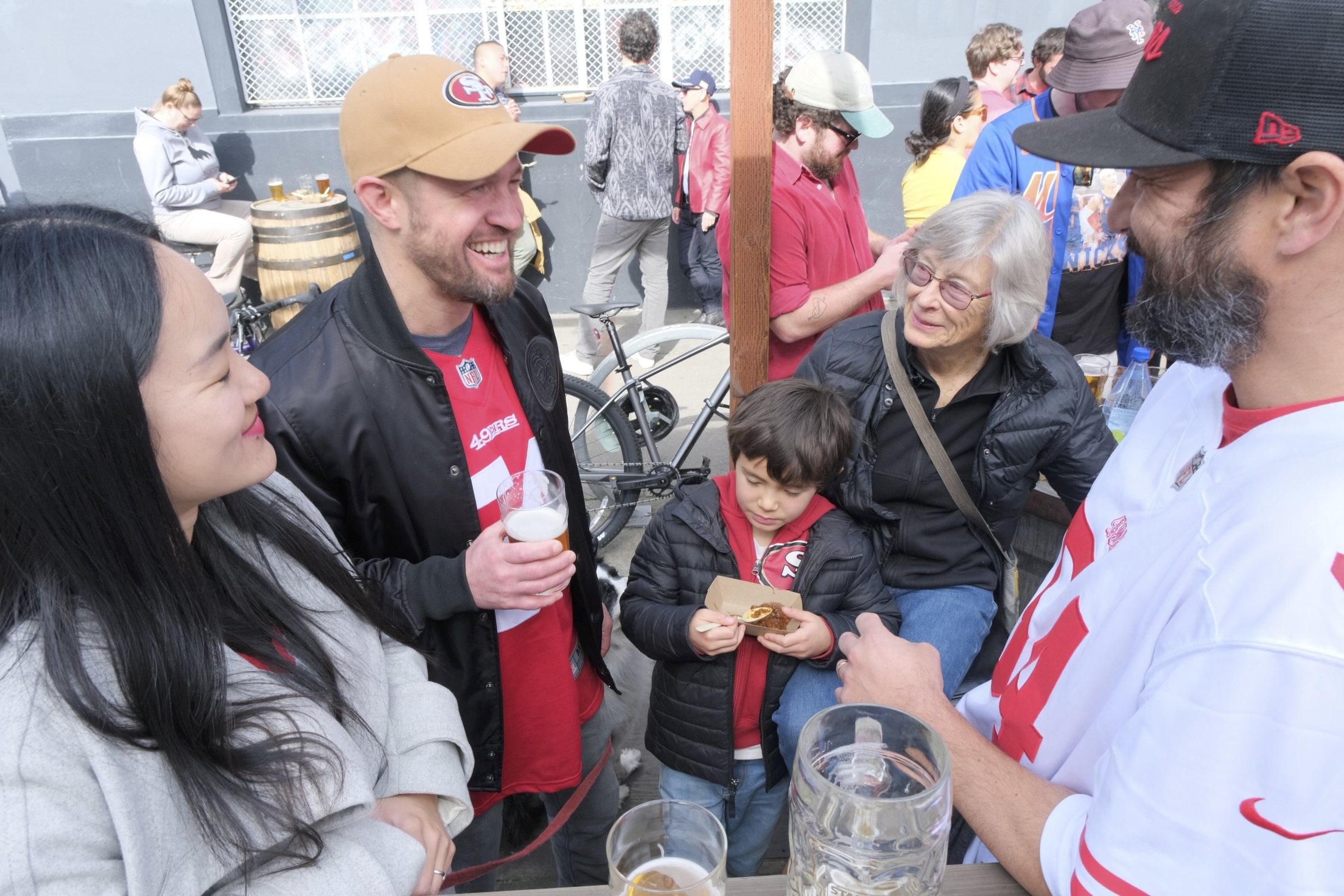 A group of people, some in sports attire, socializing outdoors with drinks; a child is focused on a snack.