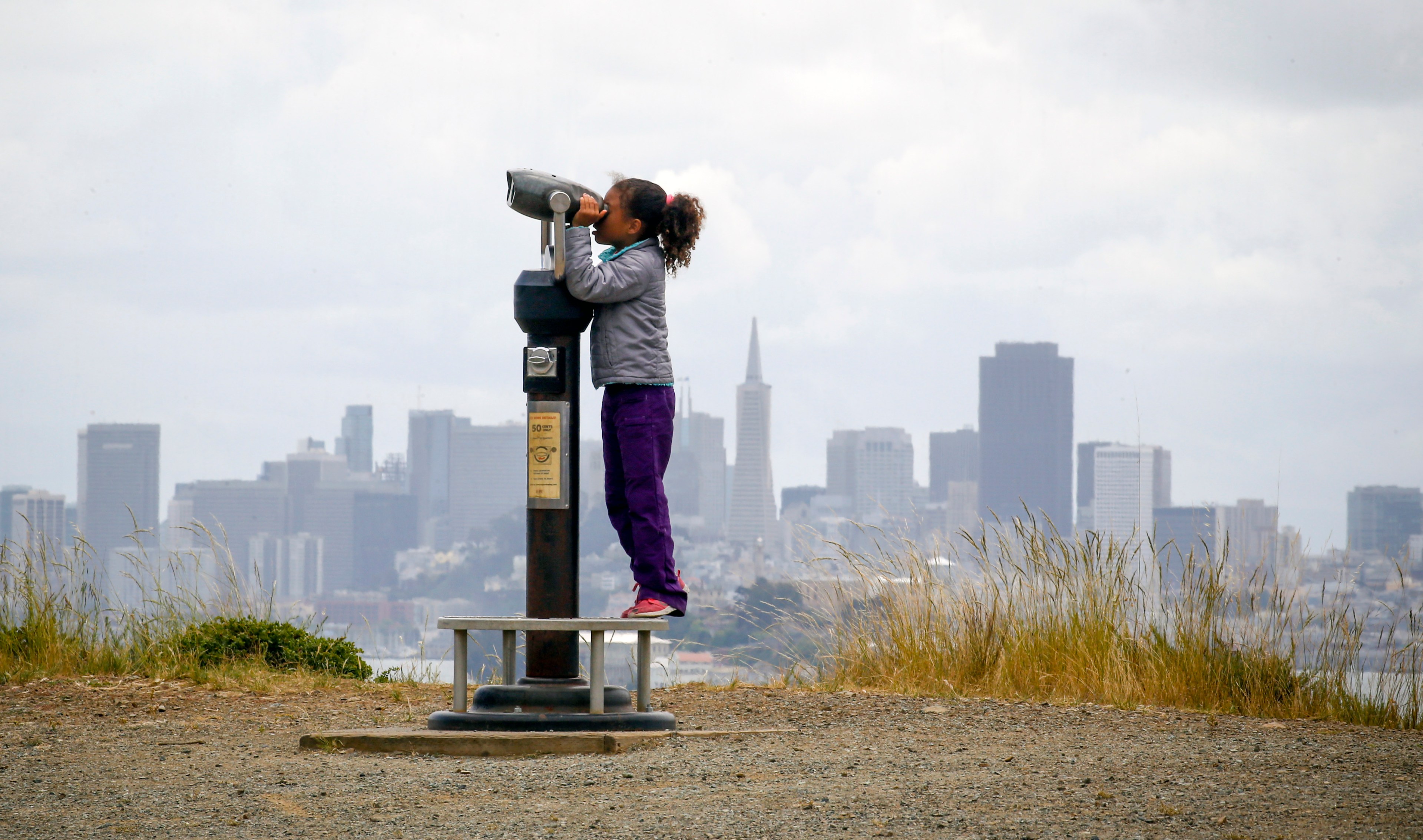 A child looks through a telescope with San Francisco skyline in the background.