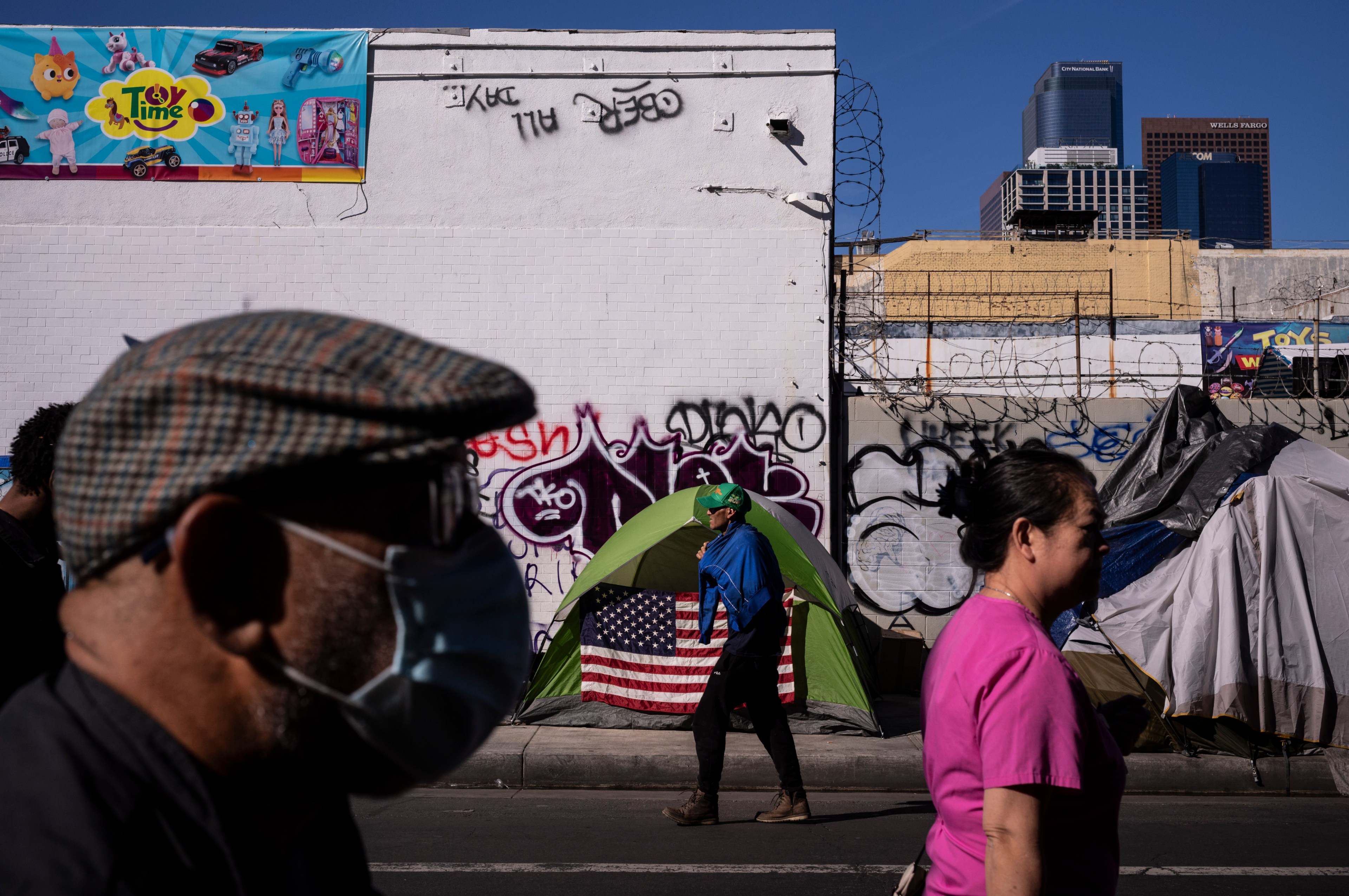 People walk by green tent with American flag set up in front of graffitied wall