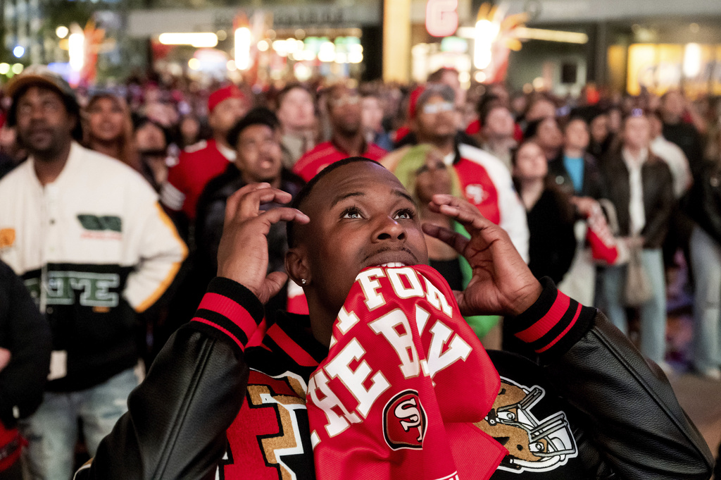A crowd looks up in anticipation; a person in the foreground in a 49ers scarf gestures excitement.