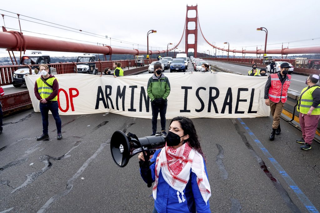 Protesters on a bridge display a banner reading "STOP ARMING ISRAEL" while a person speaks into a megaphone.