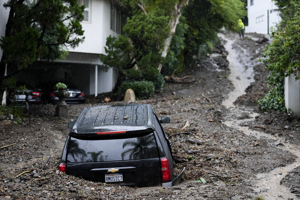 A truck is partly submerged in mud on a debris-strewn street near houses, with a person in the background.