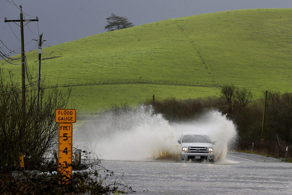 A vehicle splashes through floodwaters near a &quot;Flood Gauge&quot; sign, against a backdrop of a lush green hill.
