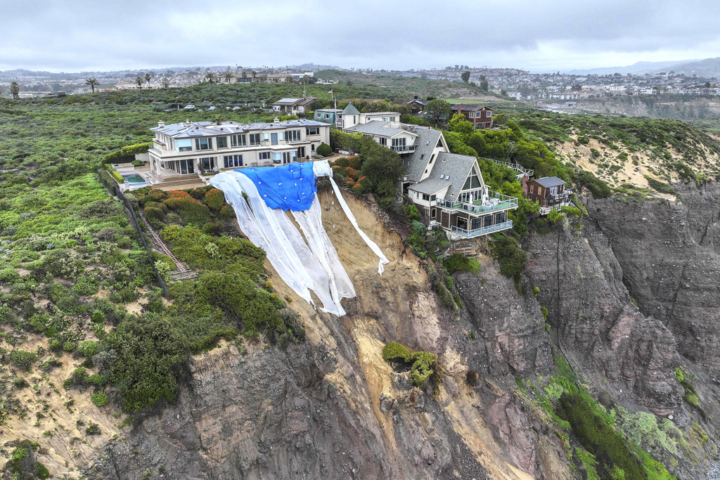 Aerial view of oceanfront homes on a cliff, one with a large blue tarp, suggesting landslide prevention.