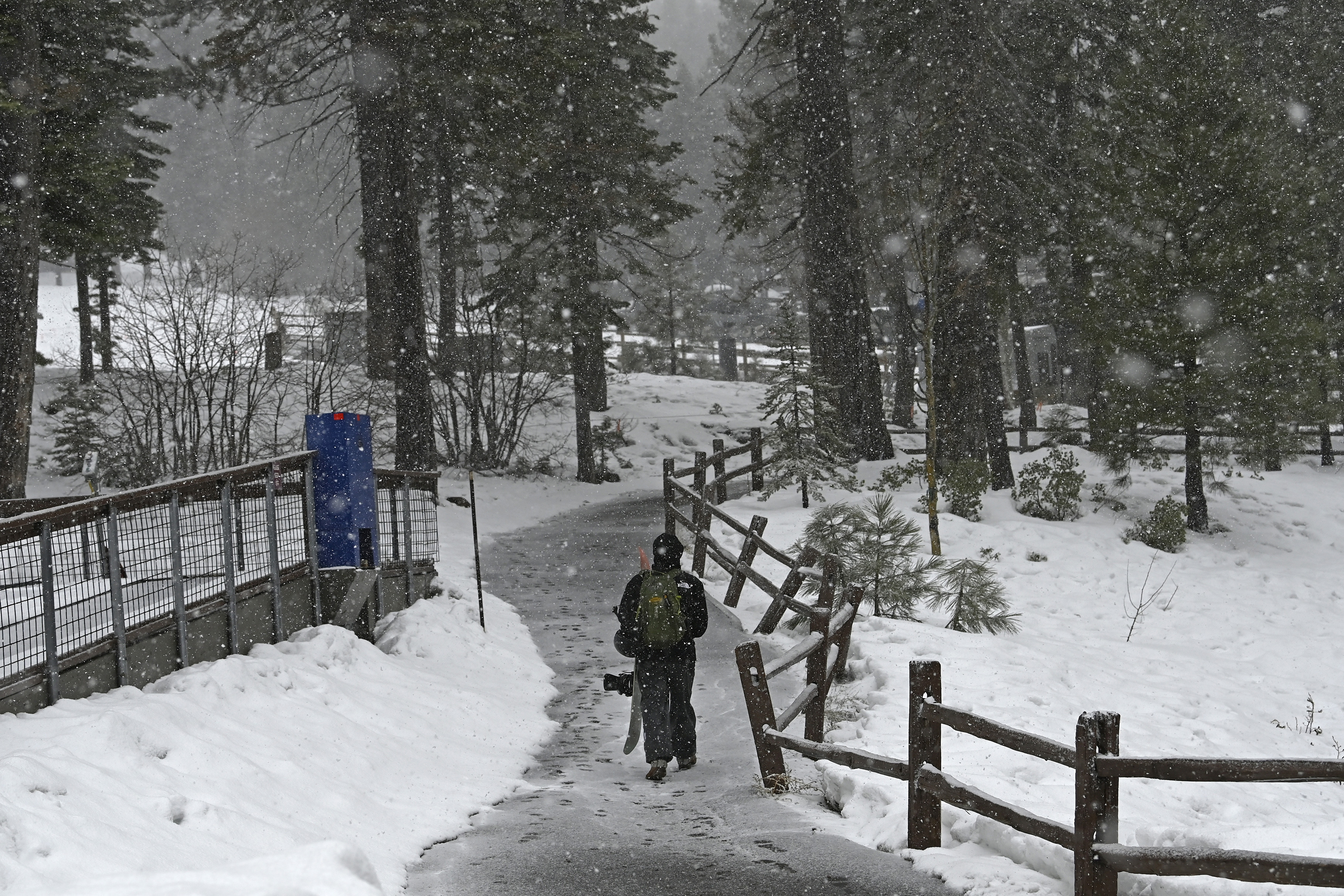A person carrying a snowboard walks down a path as snow falls
