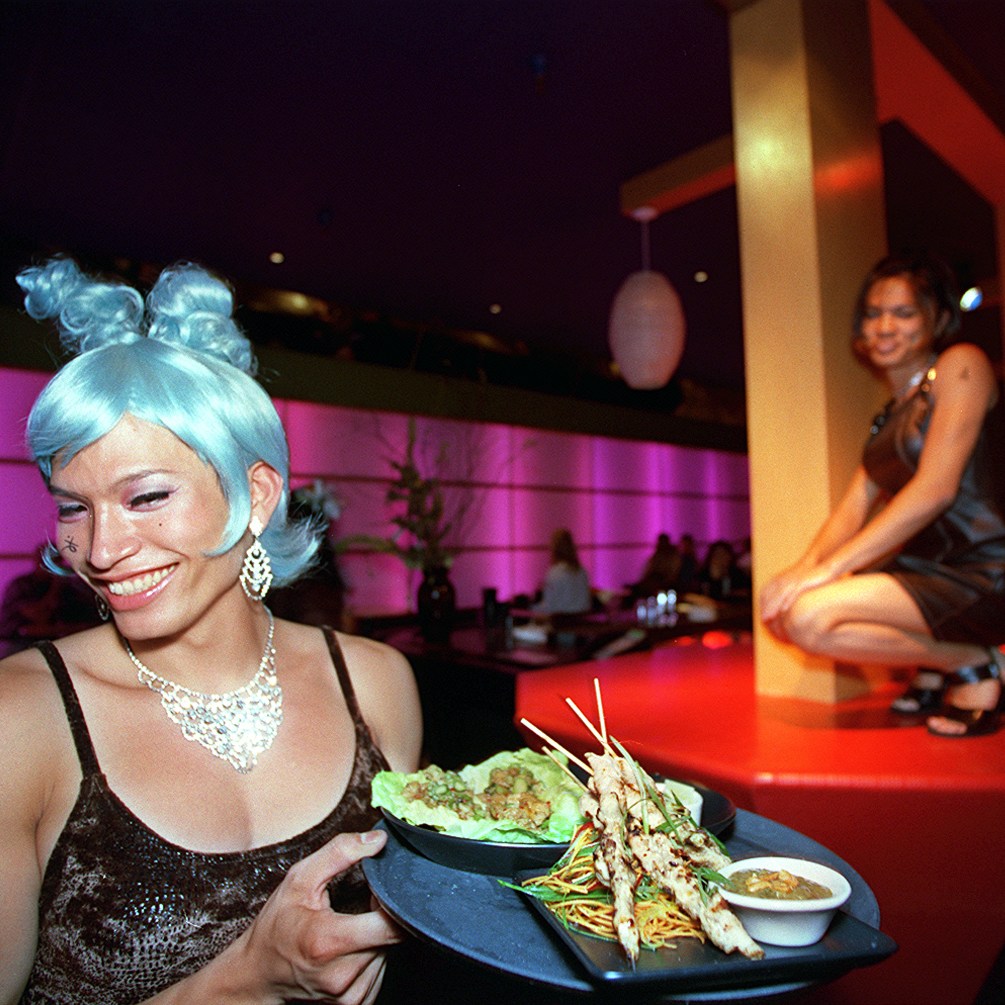 A smiling server with blue hair presents a dish; in the background, a person crouches on a lit platform in a dimly lit room.