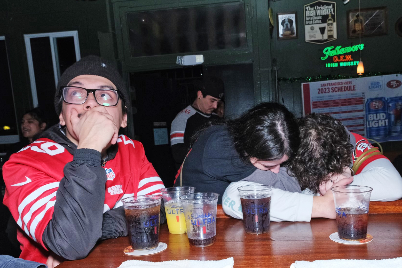 A tense moment at a sports bar: someone in a jersey looks shocked, while two others huddle, heads down. Drinks on the table suggest a social event.