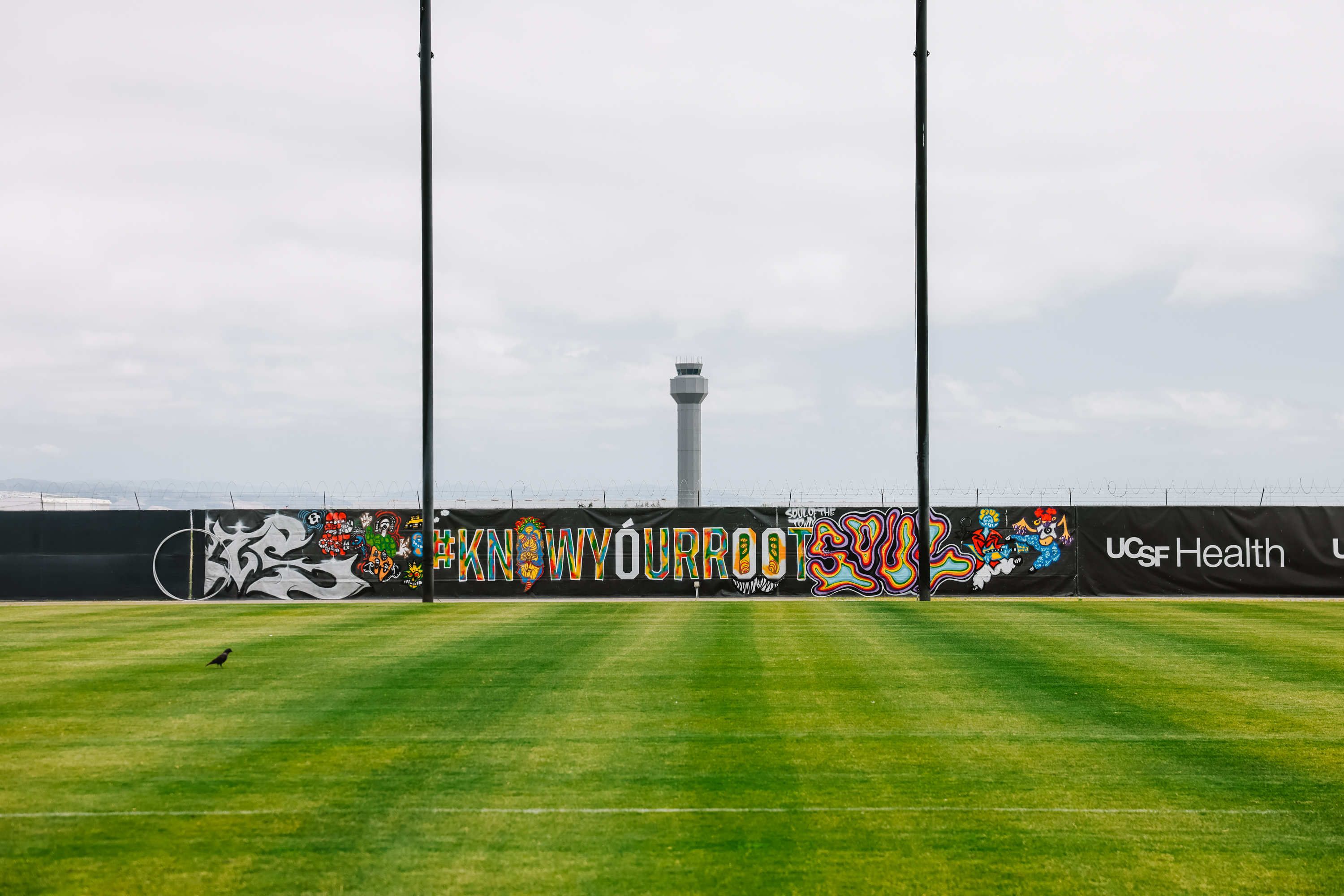 A grassy area with a colorful graffiti wall in the background, featuring the phrase &quot;#KNOWYOURROOTS&quot;, near an airport tower.
