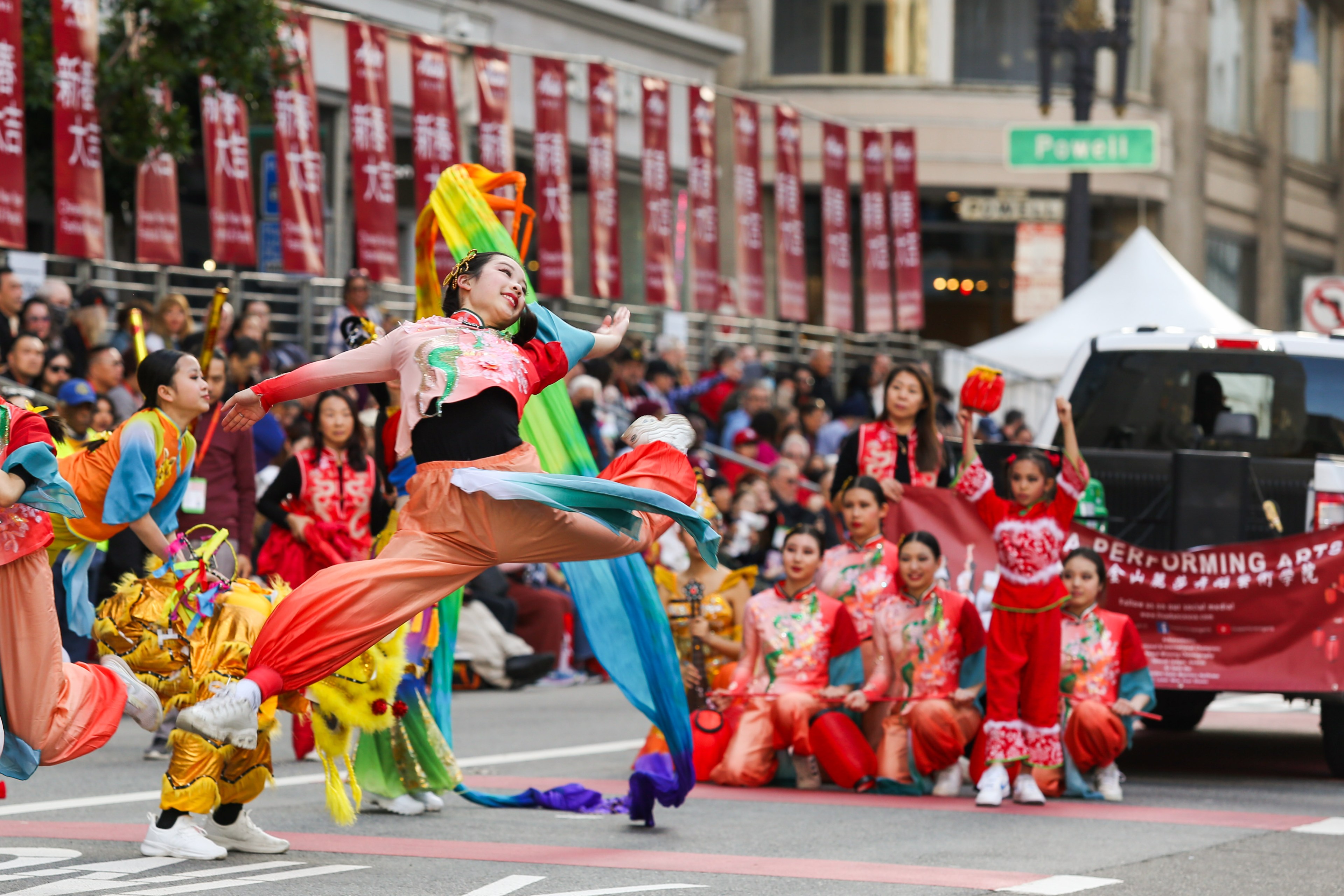 A dancer in colorful attire performs at a street parade with onlookers and participants in traditional Asian costumes.