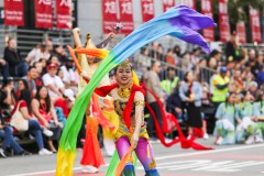 A performer in vibrant attire twirls colorful ribbons during a festive street parade.