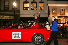 A woman waving from a red convertible with a &quot;Grand Marshal&quot; sign during a nighttime parade.