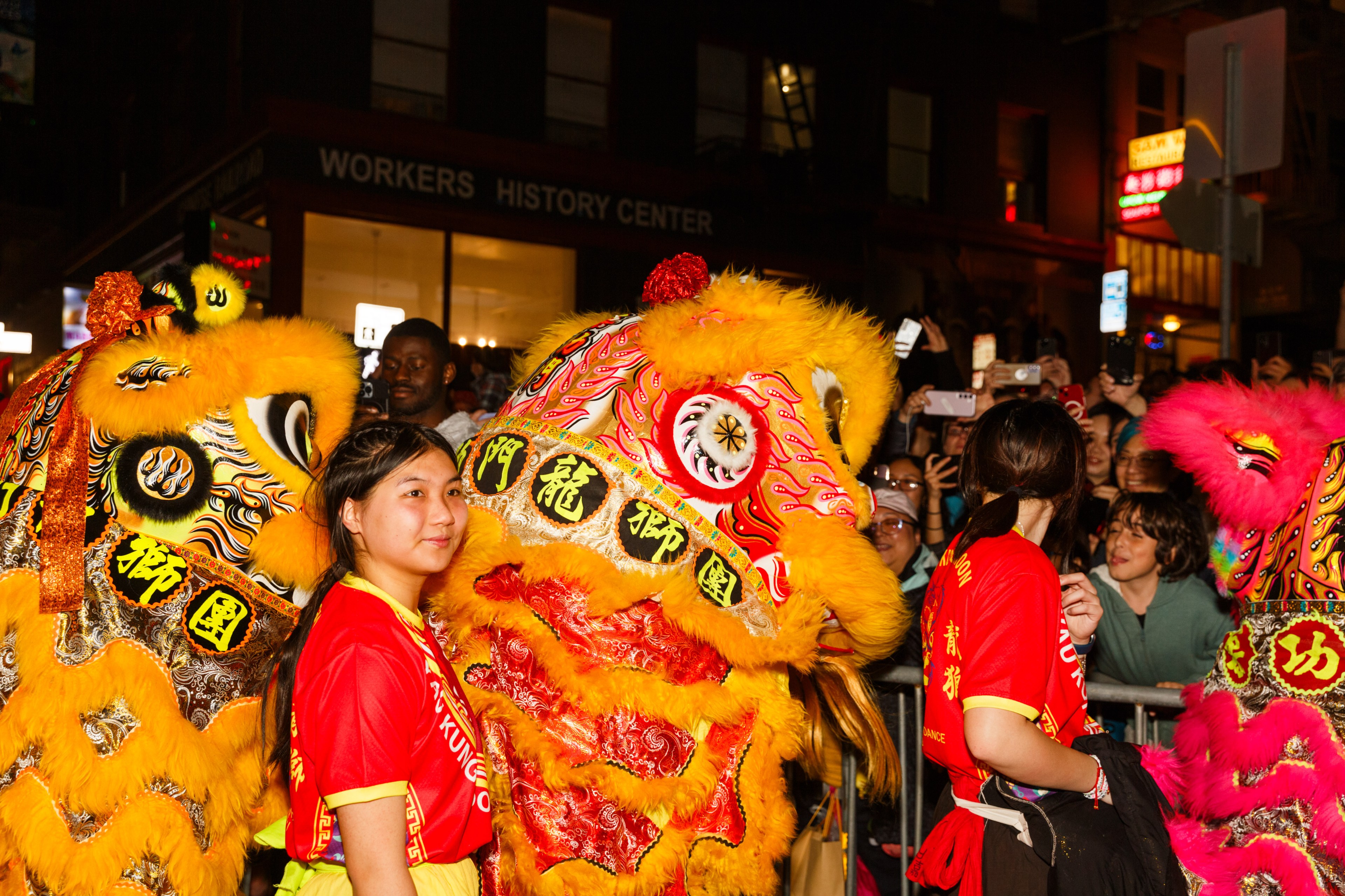 People in red shirts participate in a lion dance, surrounded by a crowd at night.