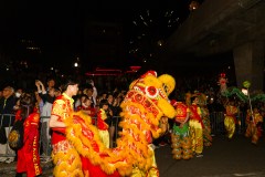 A vibrant night parade with a yellow lion dance costume, performers in red and gold attire, and a crowd watching excitedly.