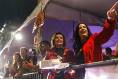 Two women in red jackets appear cheerful at an event, one pointing excitedly, surrounded by onlookers with cameras.