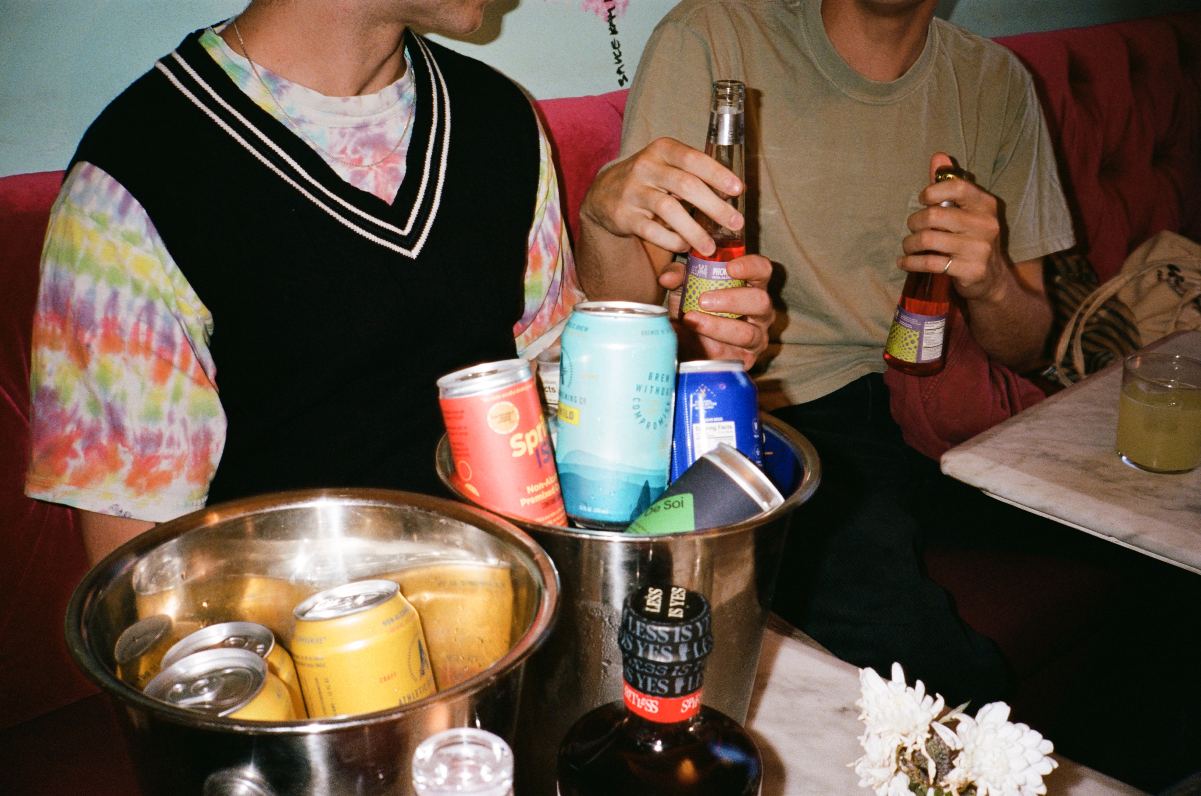 Two people are holding drinks with an ice bucket full of canned beverages on a table, along with small flowers.