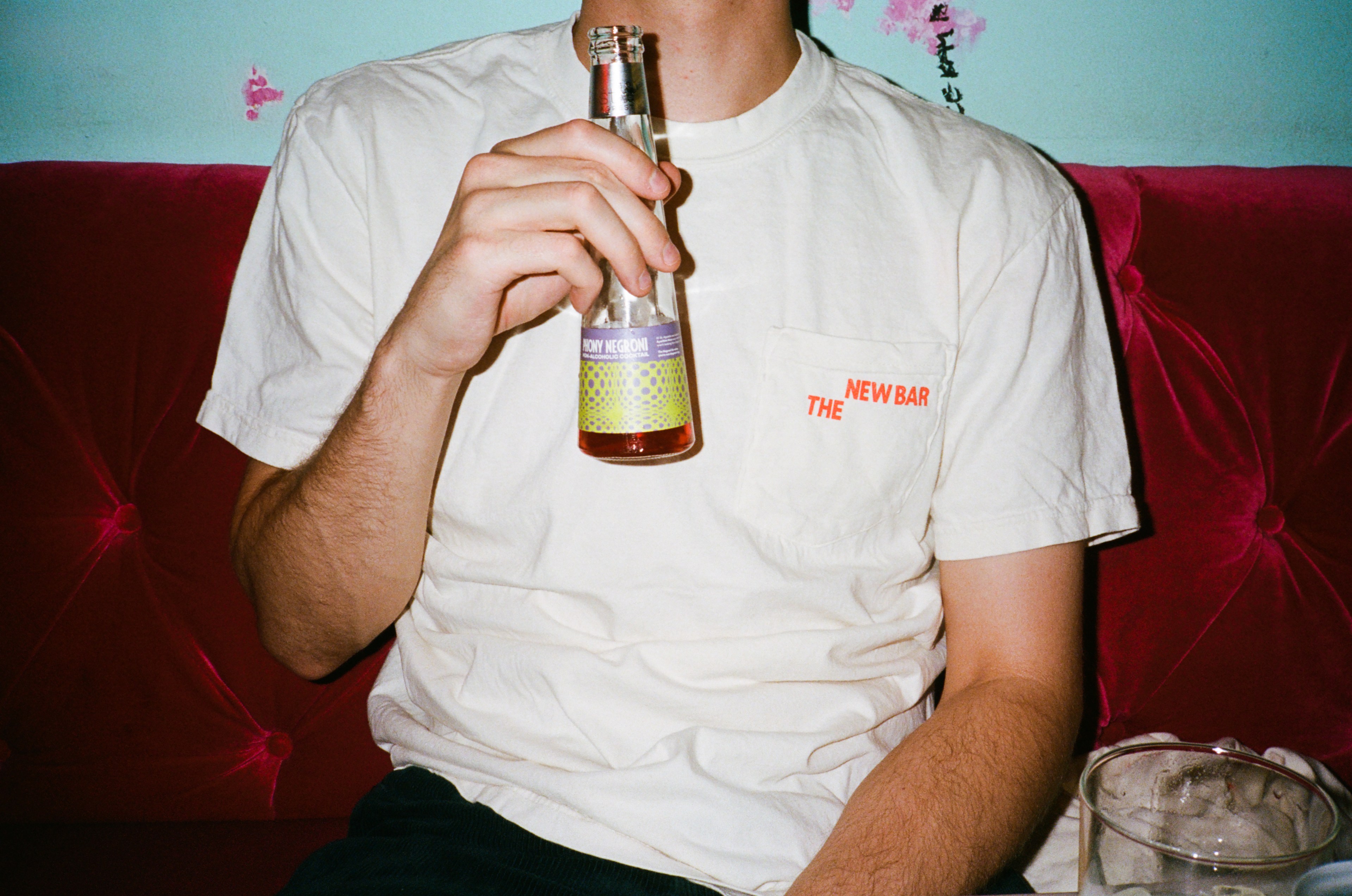 A person in a white T-shirt with the word &quot;THE NEW BAR&quot; holds a bottled drink with text &quot;HONEY NEGRONI&quot; on a red sofa.