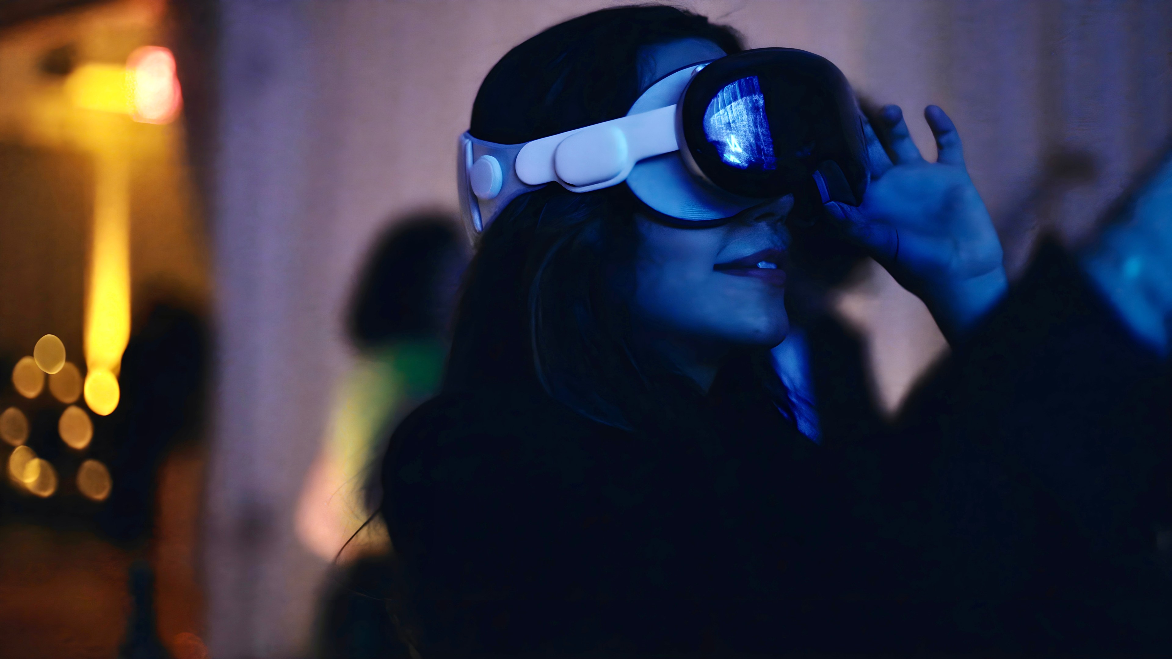 A woman wears an Apple Vision Pro headset, illuminated by glowing blue light, at a party.