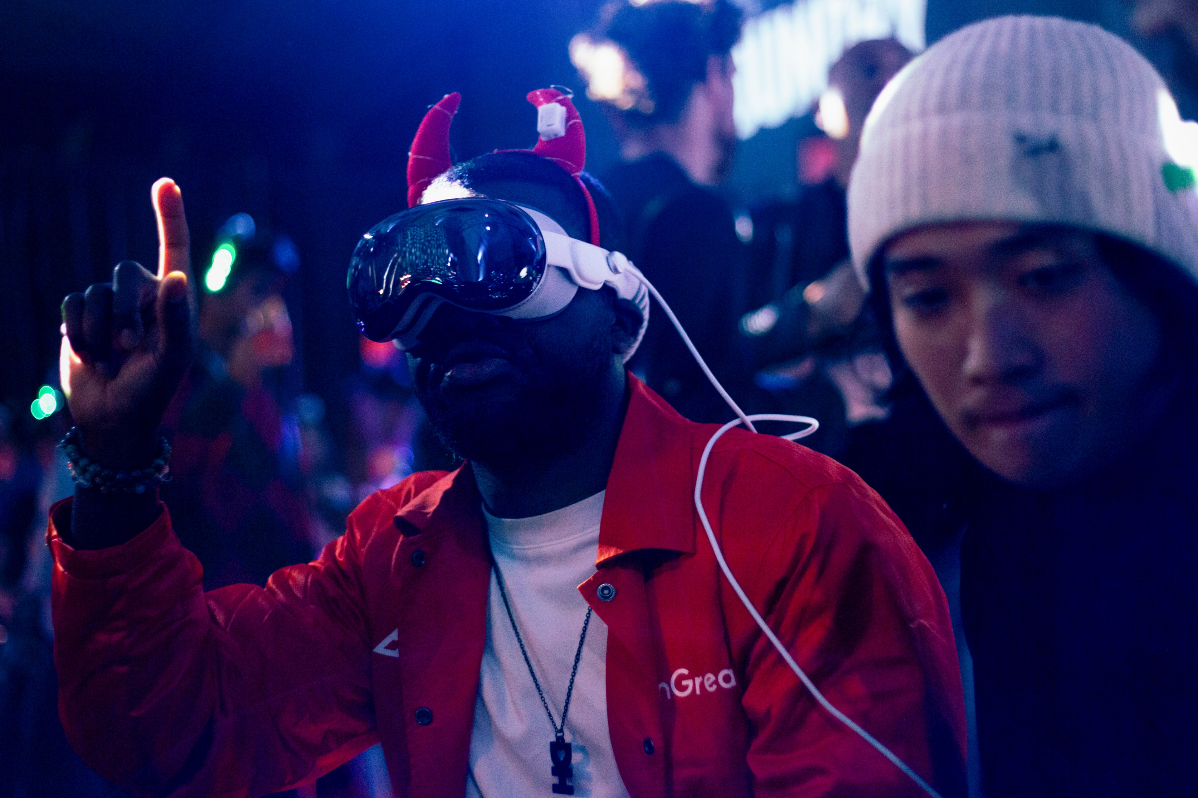 A person in a red jacket and Apple Vision Pro headset wears red cat ears and gestures with his finger up in the air next to another person.