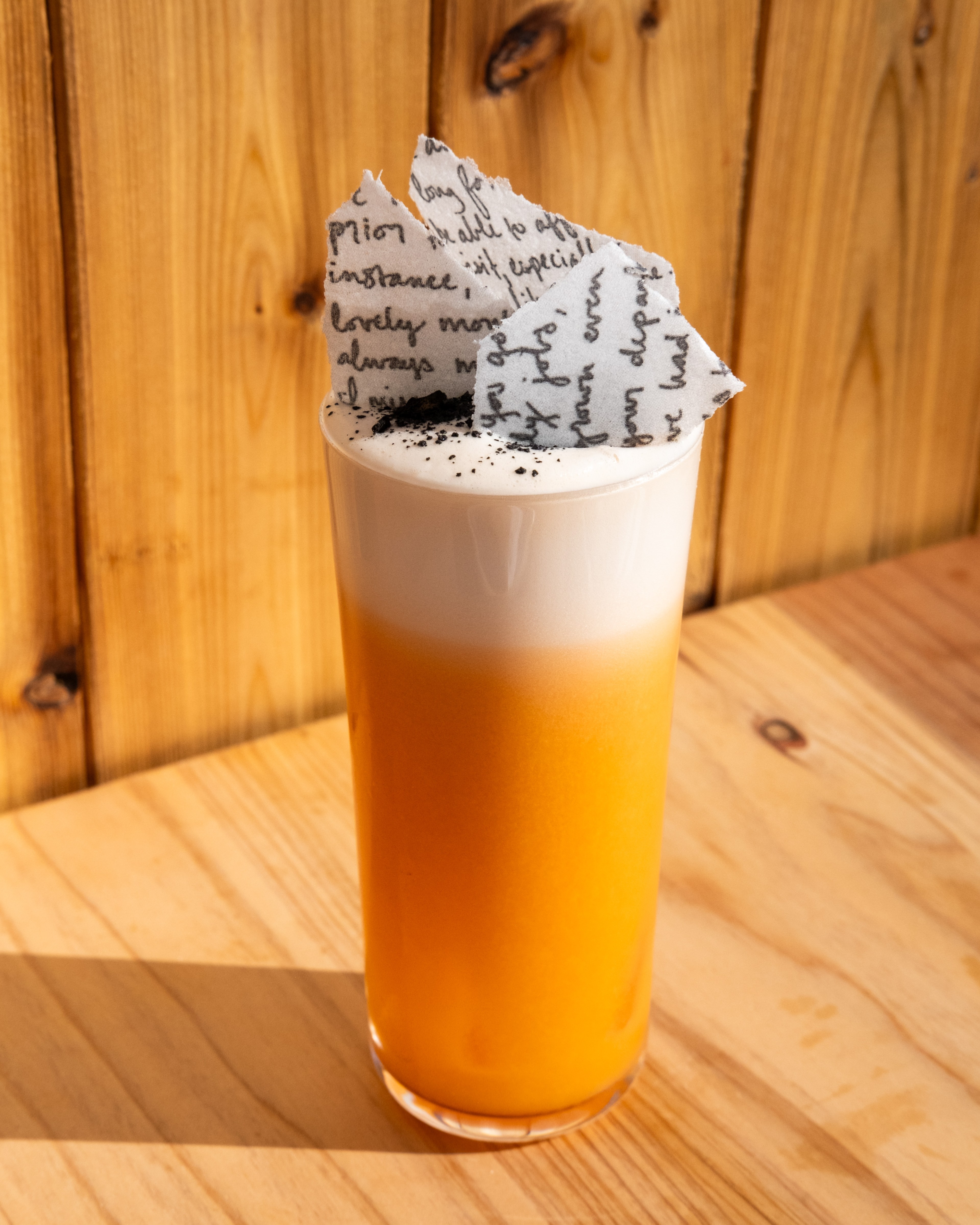 An orange cocktail in a tall glass is topped by pieces of a burnt rice paper love note.