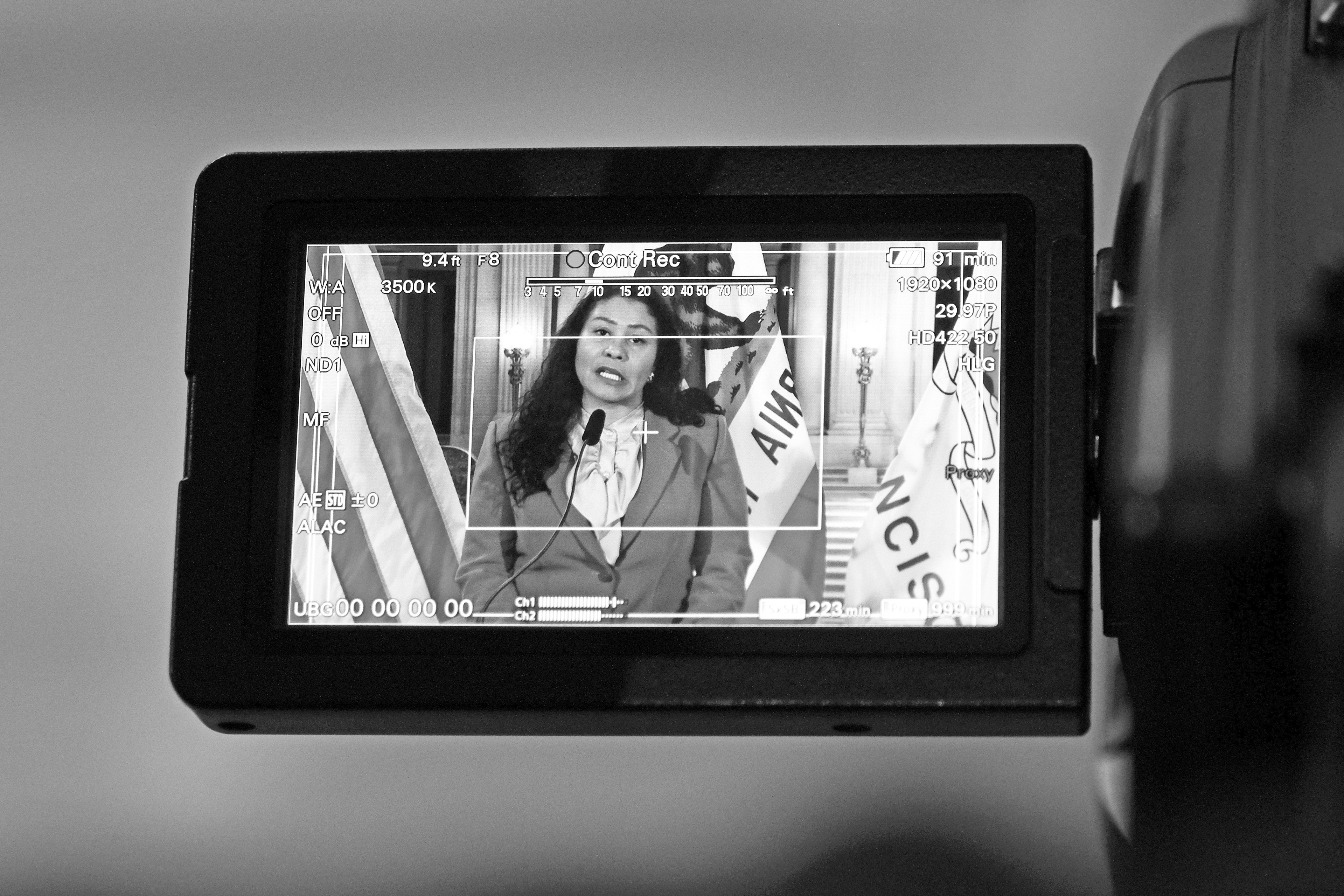 A camera's LCD screen shows San Francisco Mayor London Breed speaking, with camera settings overlaid, all in grayscale.