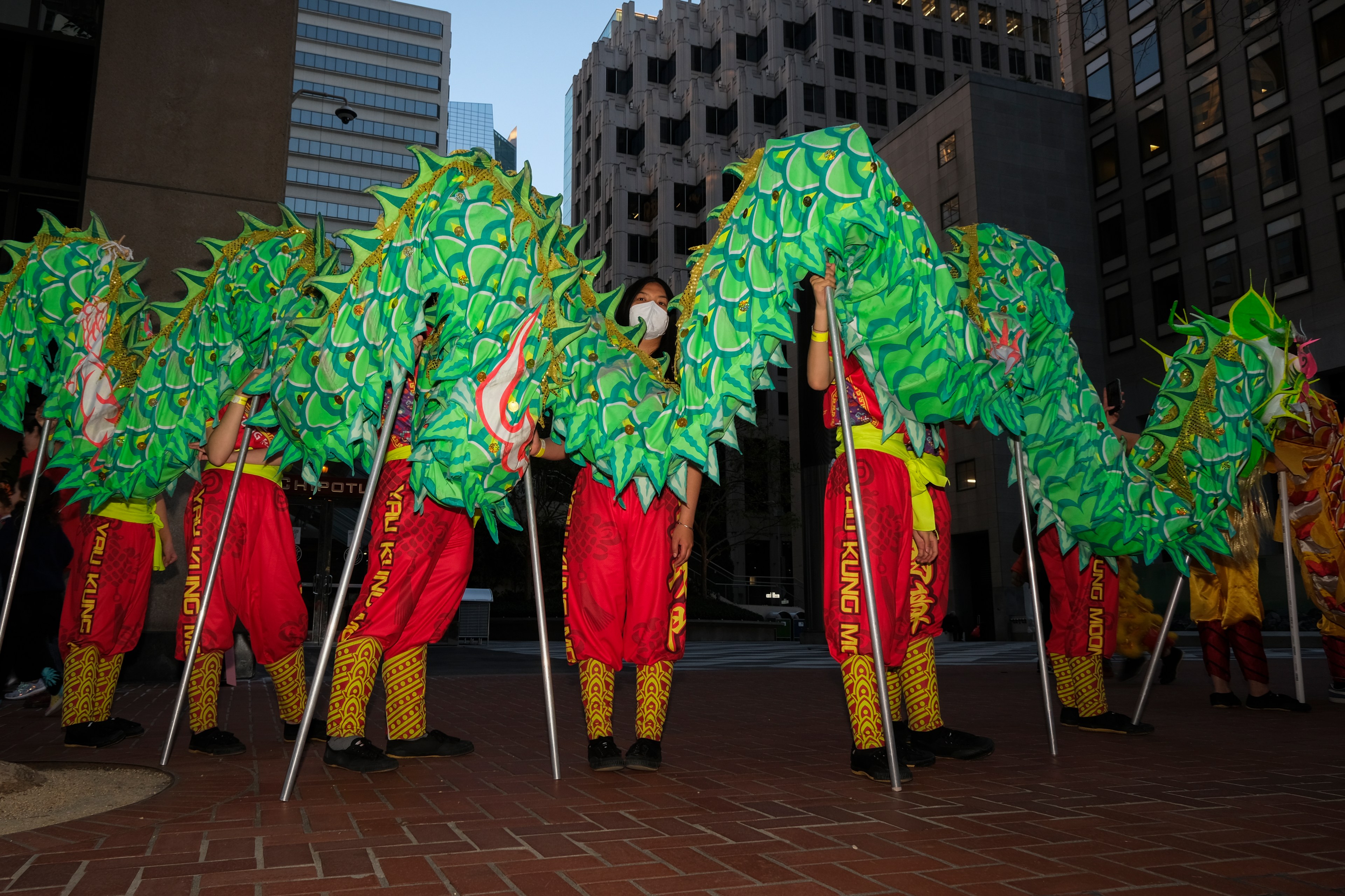 Performers in red and gold holding a long, green dragon costume, parading against an urban backdrop.