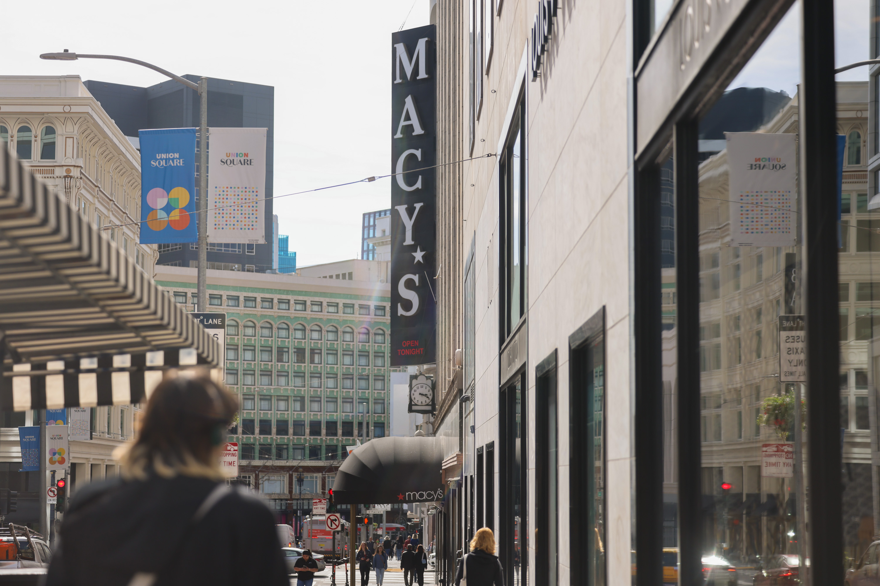 A city street with Macy's signage, walking people, and buildings under a clear sky.