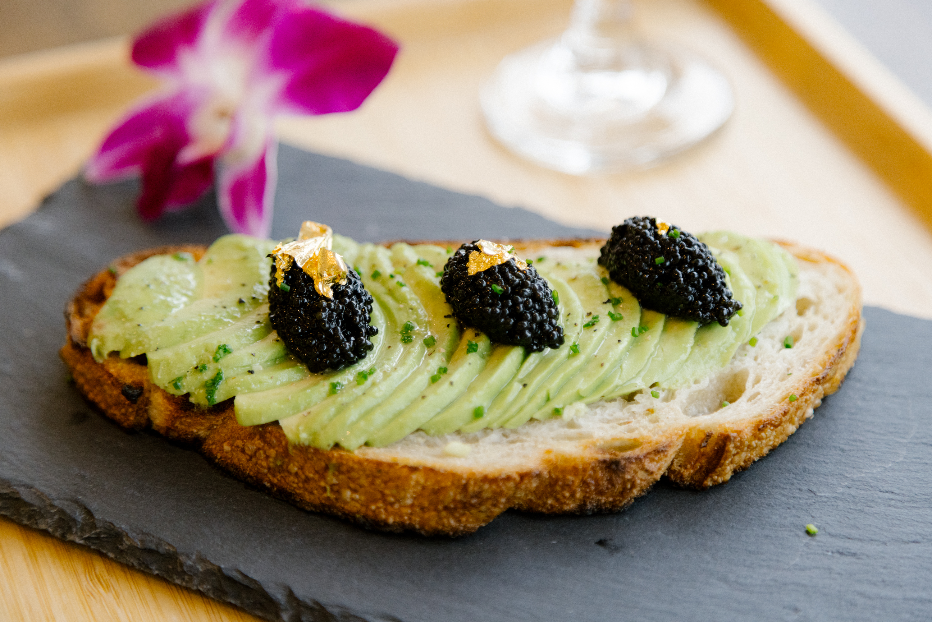 A slice of bread topped with avocado slices, caviar, and gold leaf on a slate plate, garnished with a purple flower.