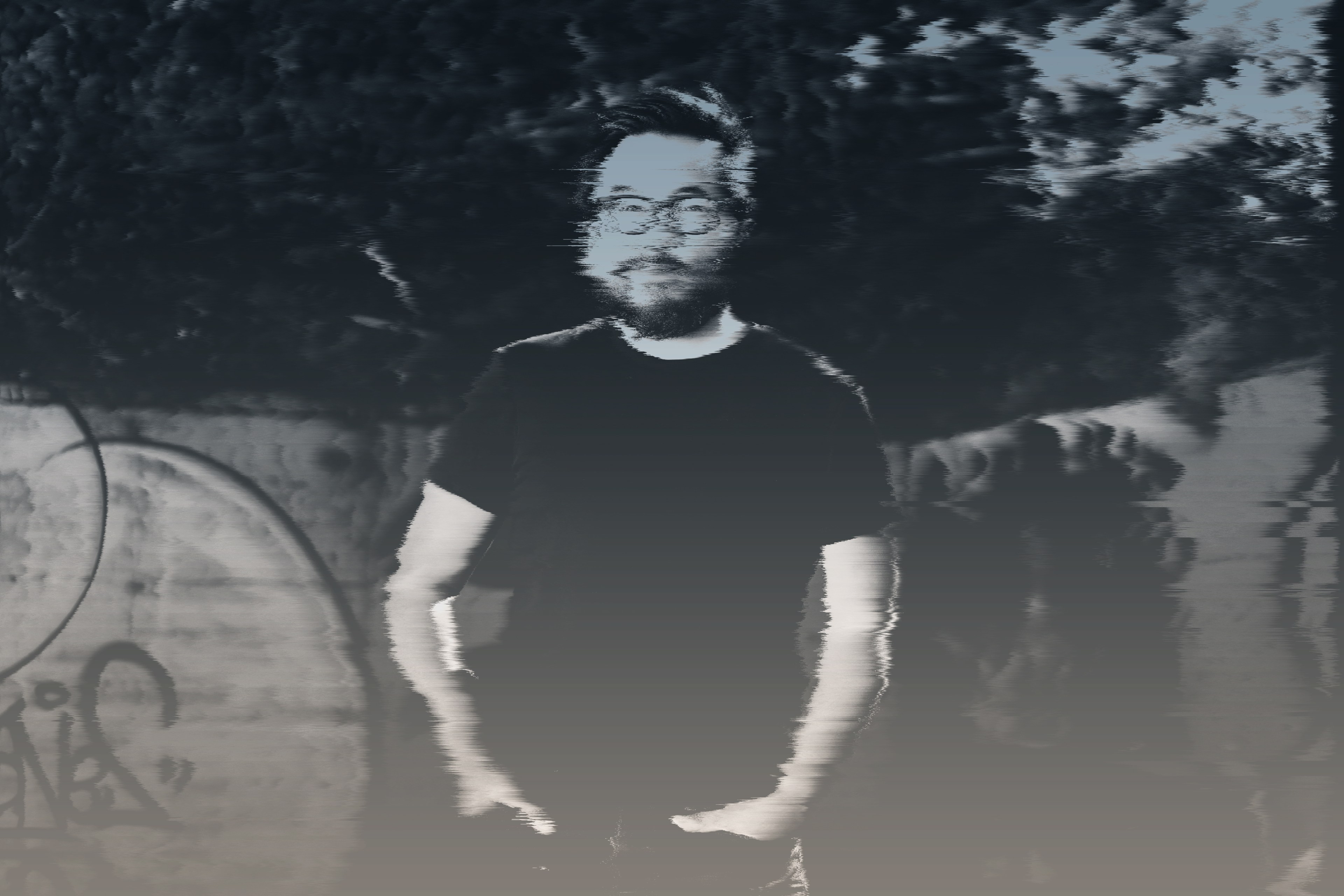 A man with glasses and a beard stands in a dim, desaturated setting with graffiti in the background. The image is grainy with a ghostly visual effect.
