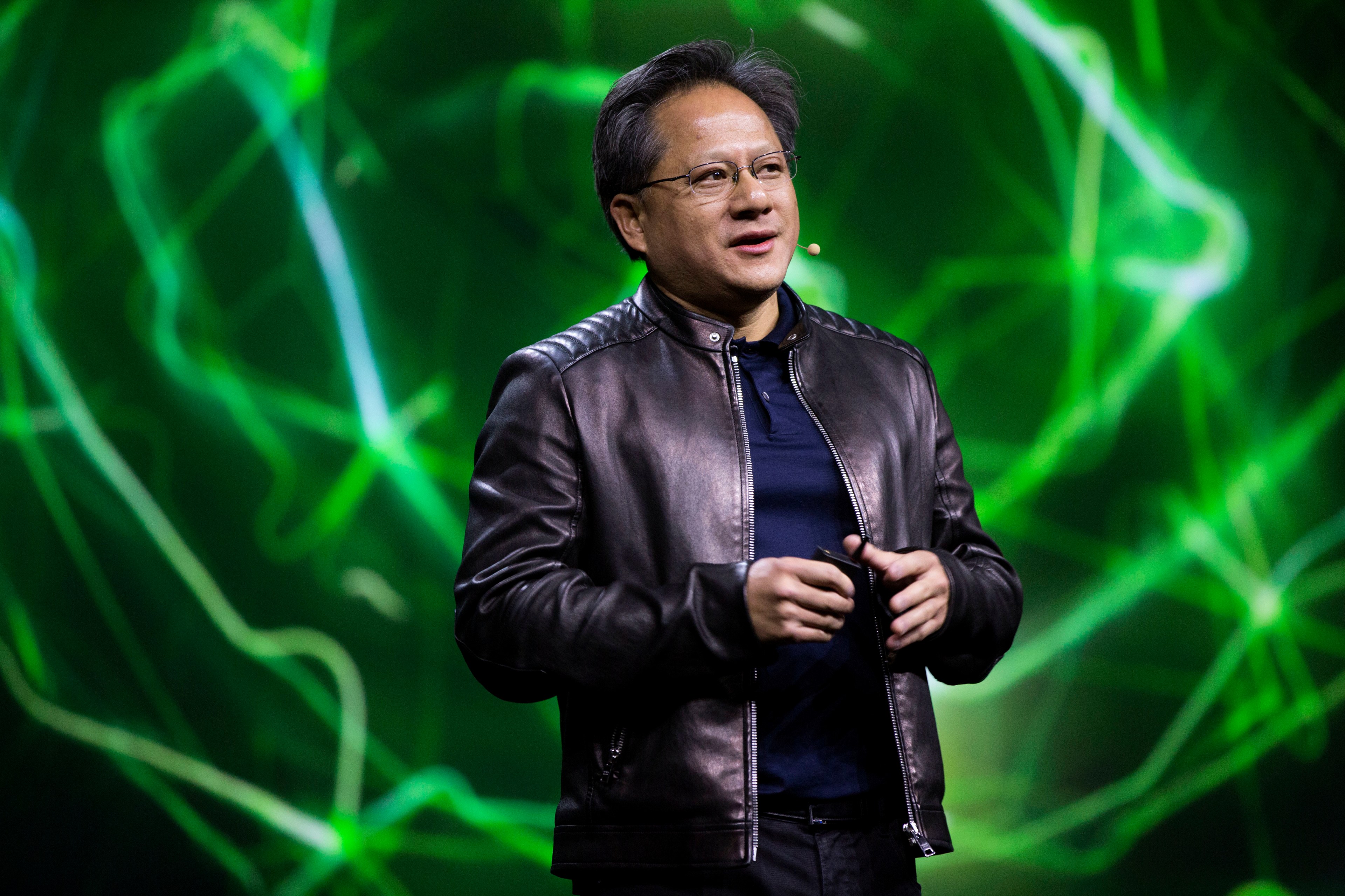 A person is speaking on stage with dynamic green lighting in the background. They're wearing glasses and a black leather jacket.
