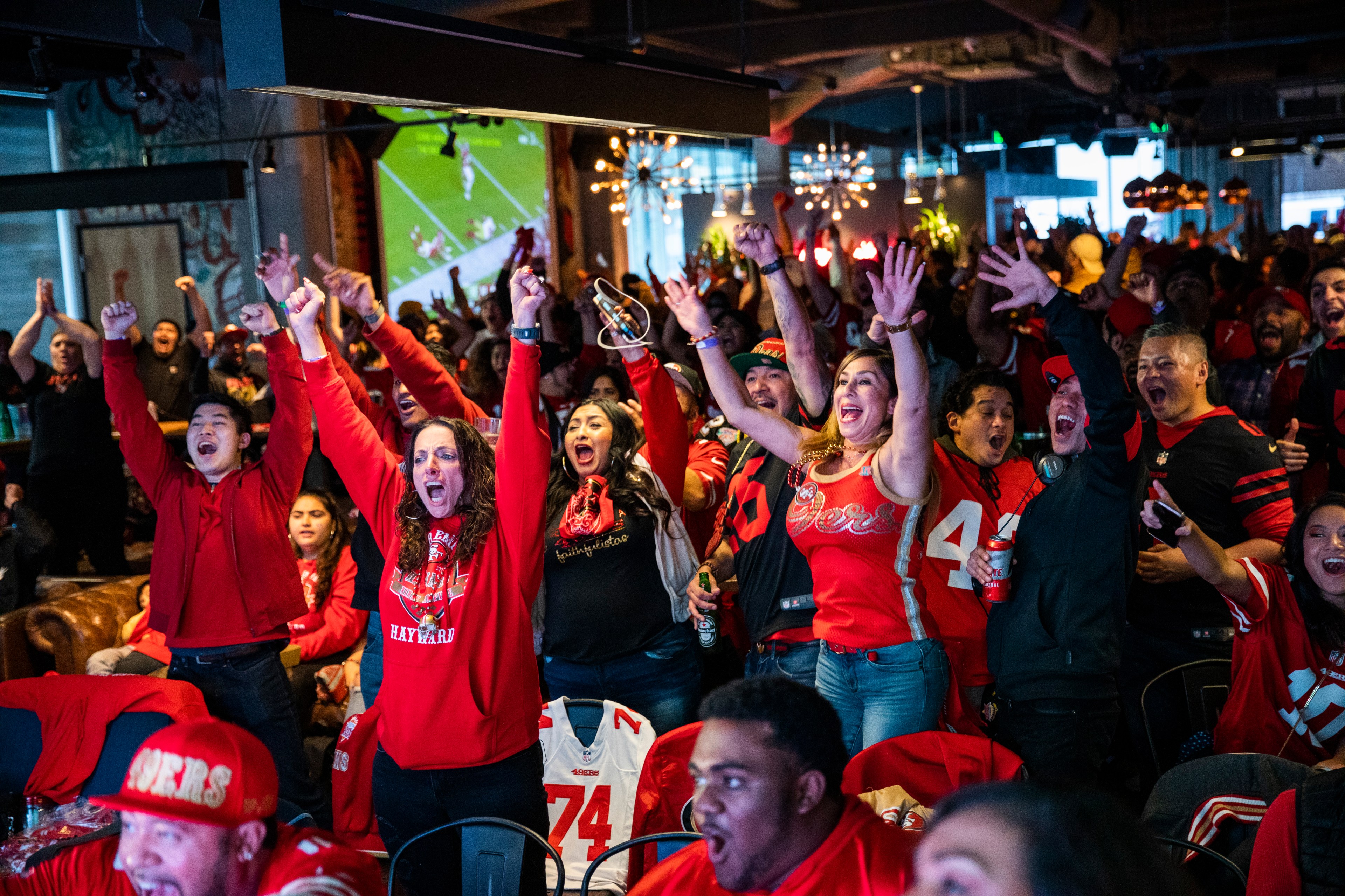A large group of people wearing San Francisco 49ers apparel jump and raise their hands in the air while celebrating inside a sports bar.