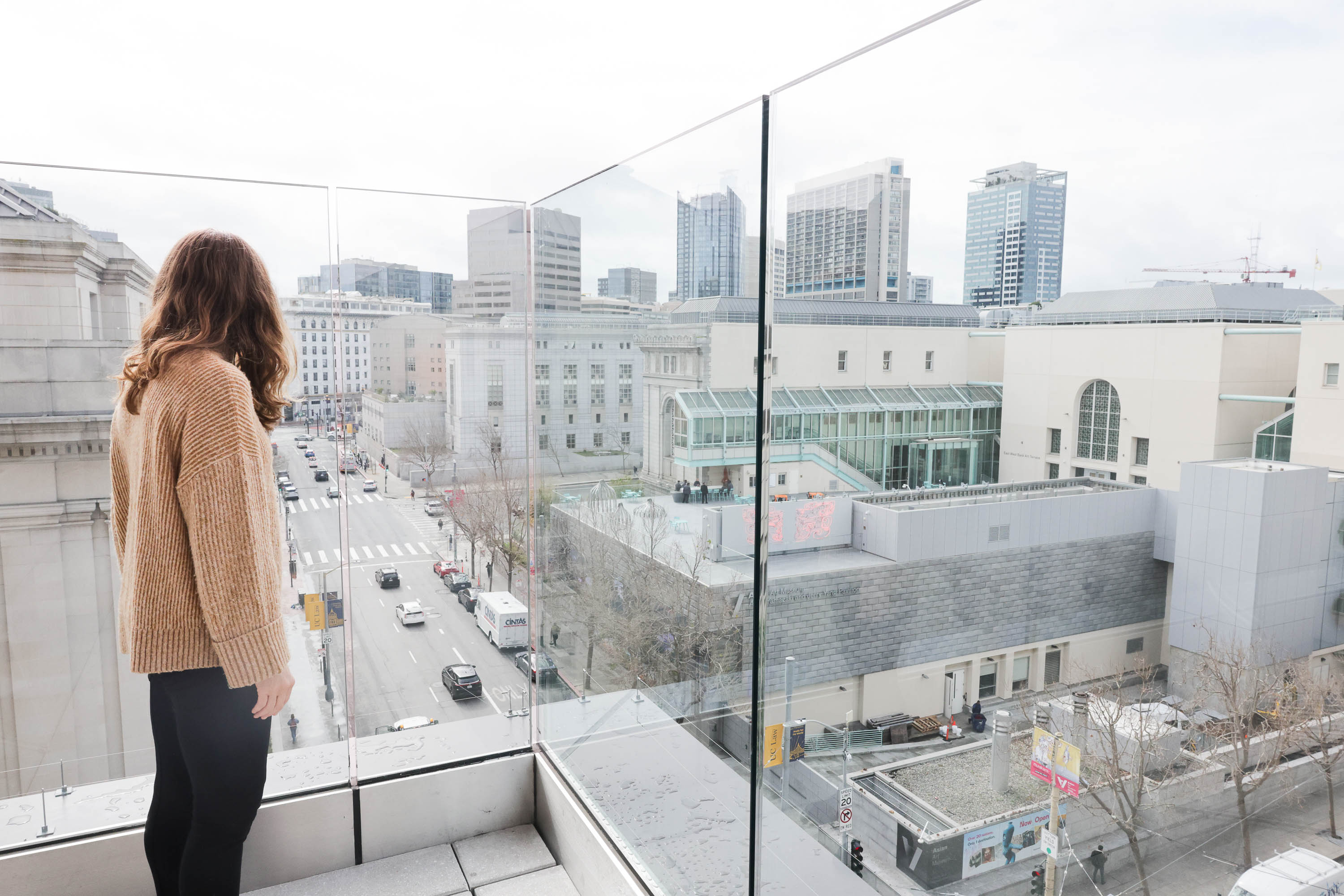 A woman views a cityscape with buildings and streets from a balcony with glass barriers.