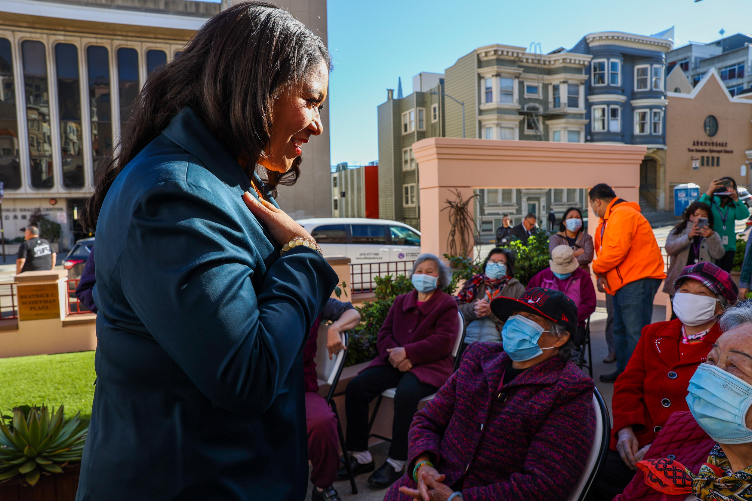 A woman gestures while speaking to seated, masked elderly individuals outdoors. Urban setting, sunny day.