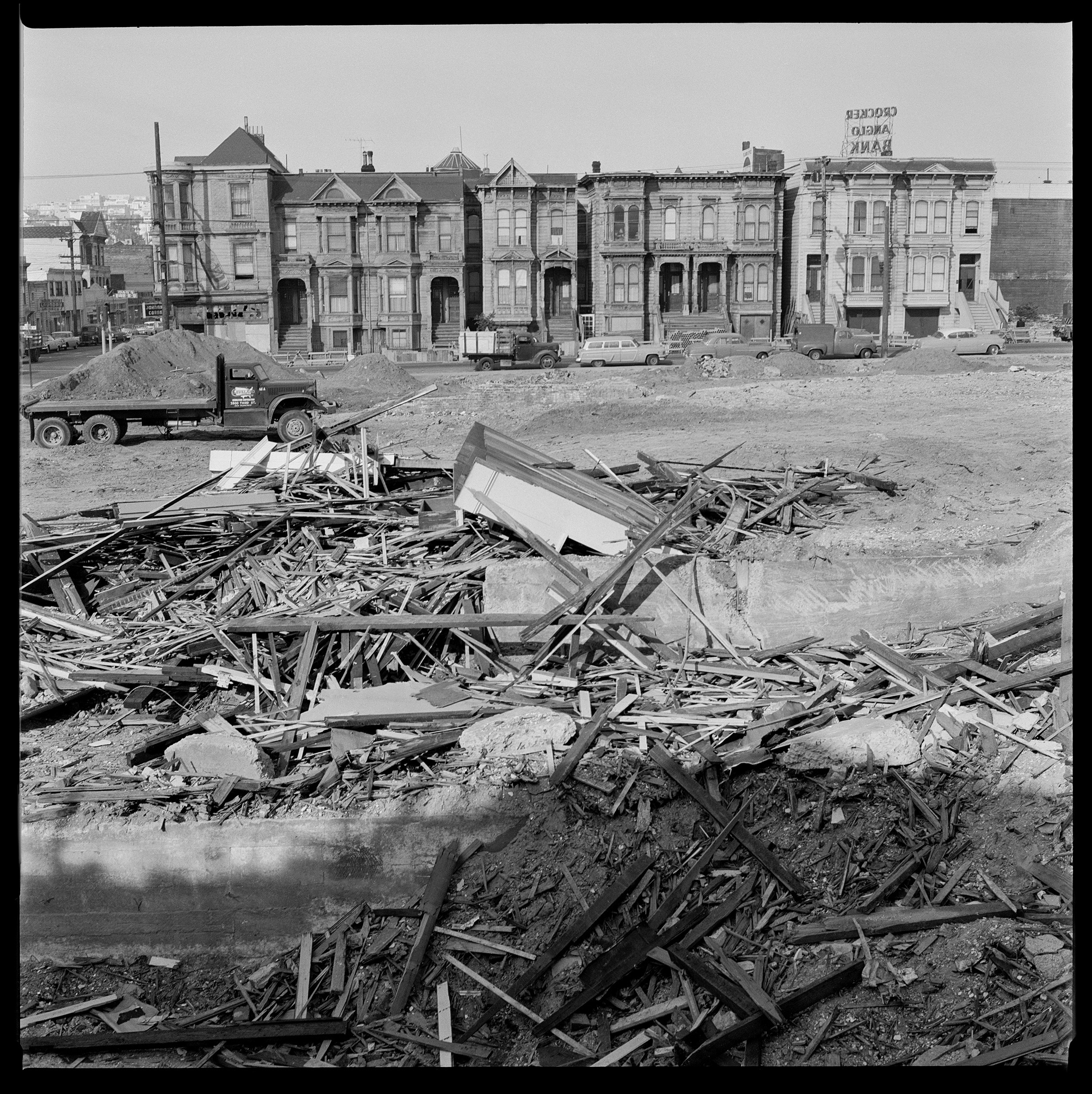 A black and white photo shows a pile of wooden debris in the foreground, with Victorian houses and a truck in the background.