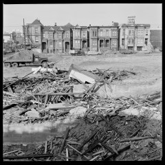 A black and white photo shows a pile of wooden debris in the foreground, with Victorian houses and a truck in the background.