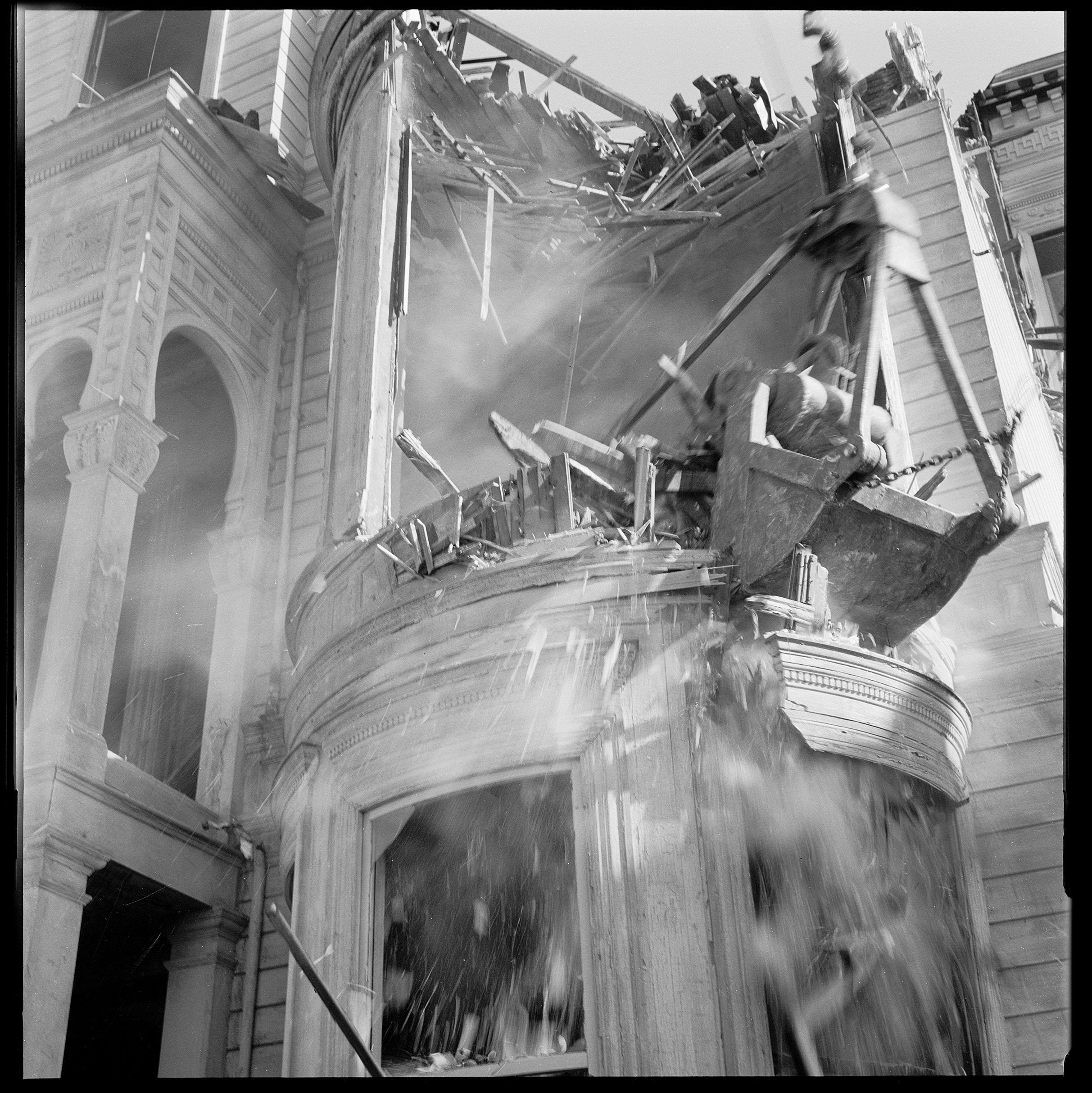 A black and white photo of a collapsing building with debris falling and water spraying upwards.