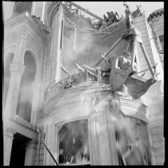 A black and white photo of a collapsing building with debris falling and water spraying upwards.