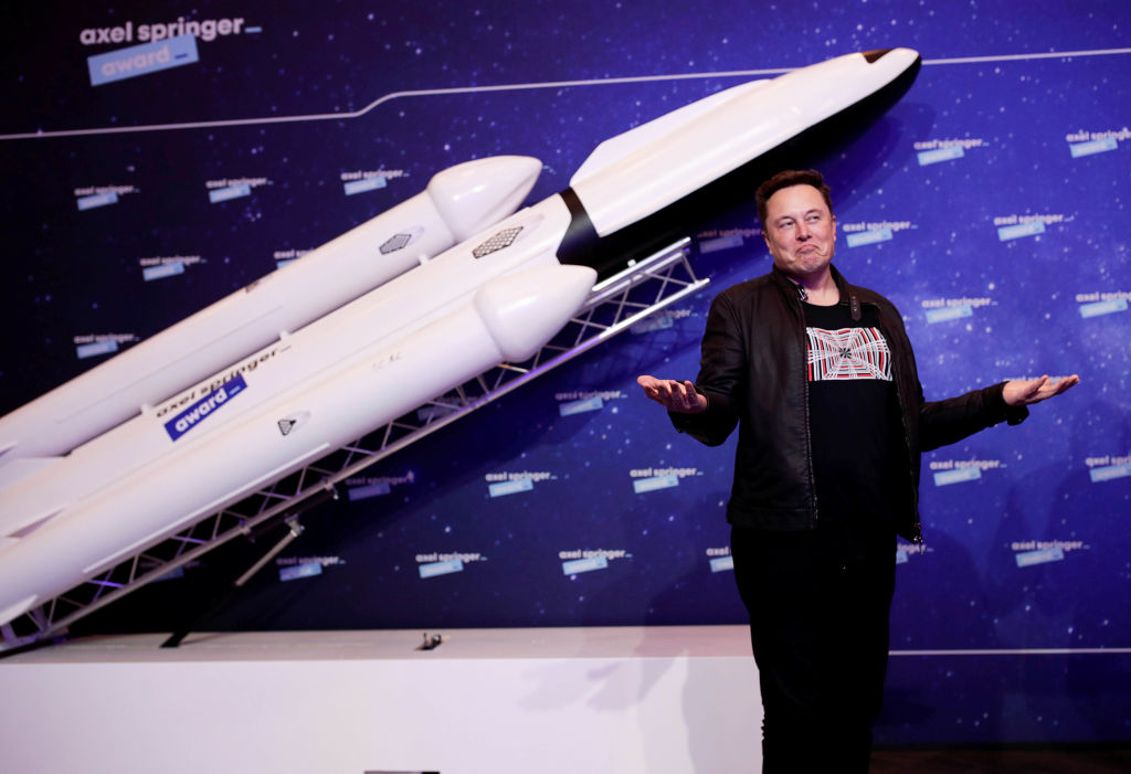 A man stands smiling with hands outstretched before a model of a white, multi-stage rocket at an event.