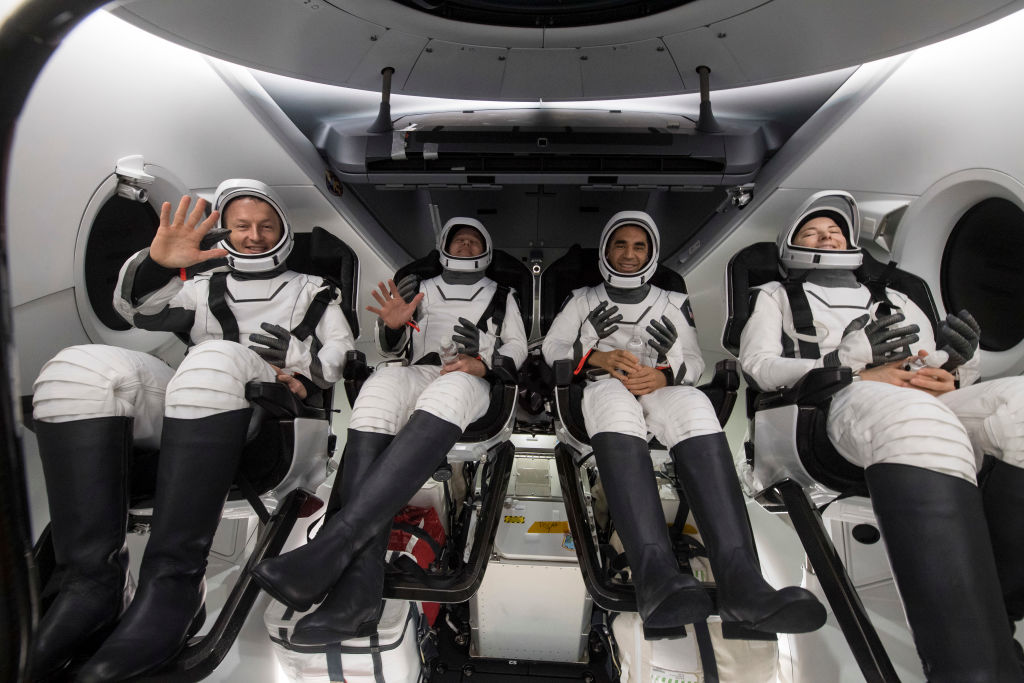 Four astronauts in white suits wave from inside a spacecraft. They're strapped in with helmets on laps.
