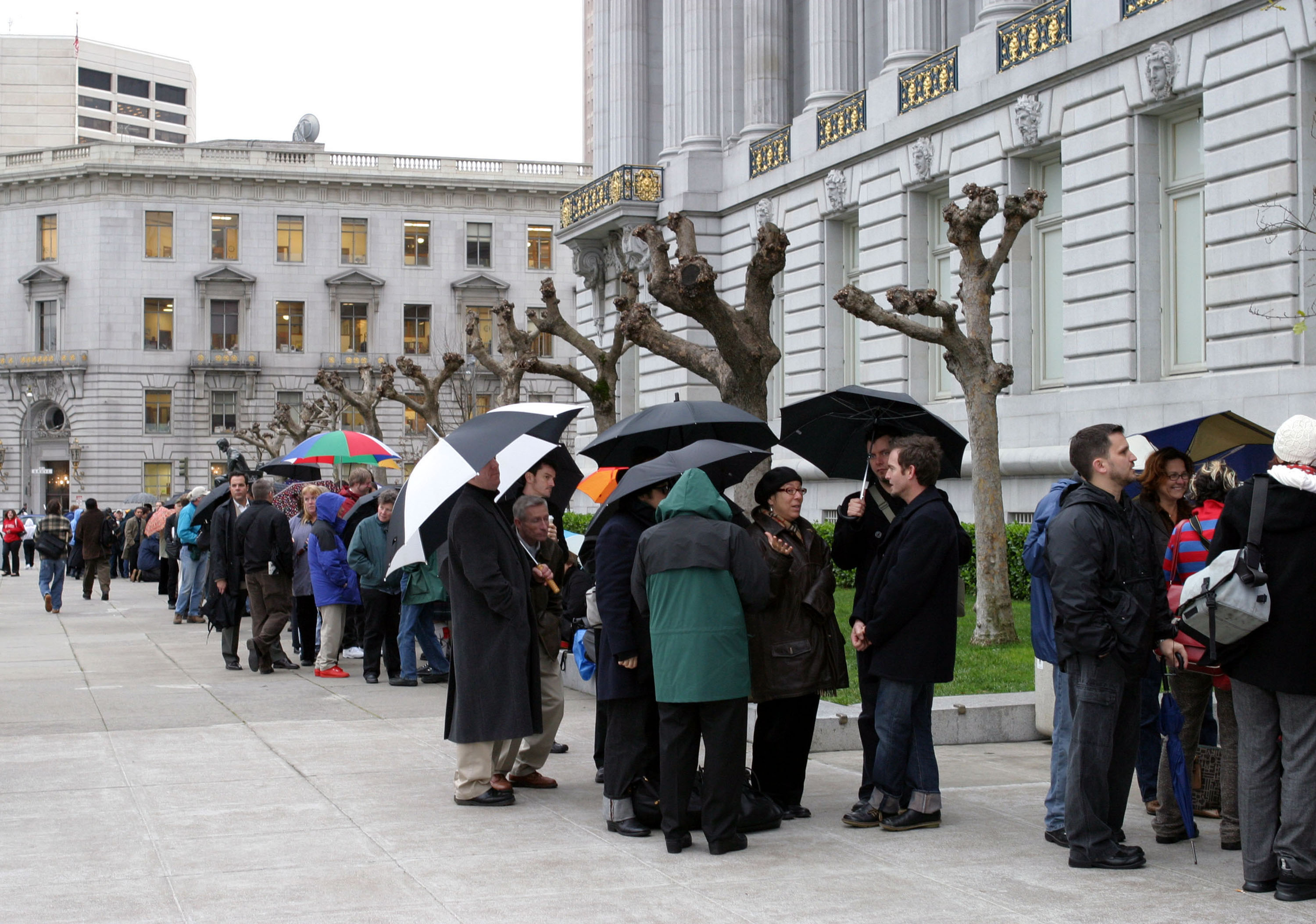 A line of people, many wearing rain coats and holding umbrellas, stands outside of San Francisco City Hall. The line stretches down the block.