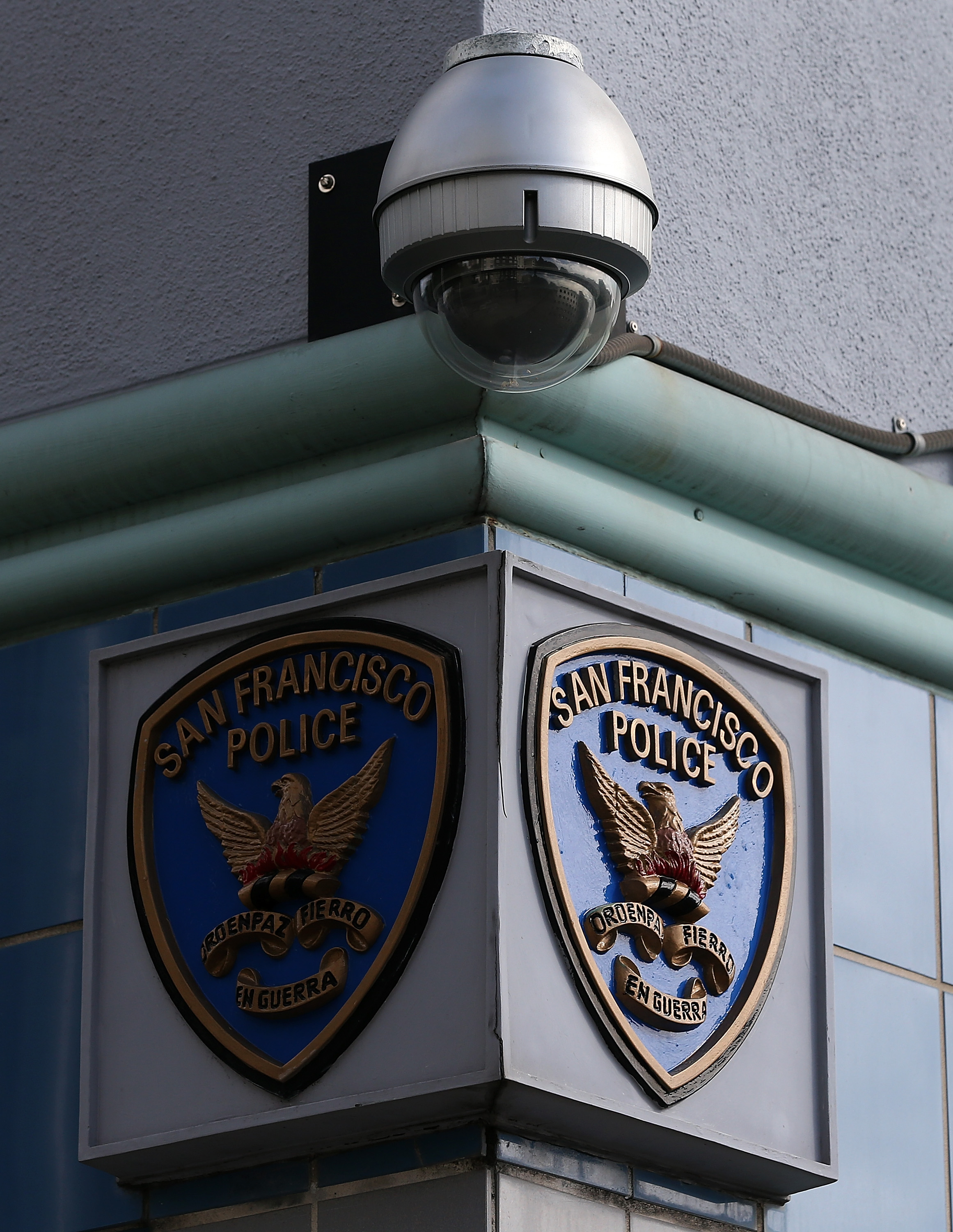 The image shows a corner of a building with two &quot;San Francisco Police&quot; shields mounted underneath a surveillance camera. The shields feature an eagle and a banner.