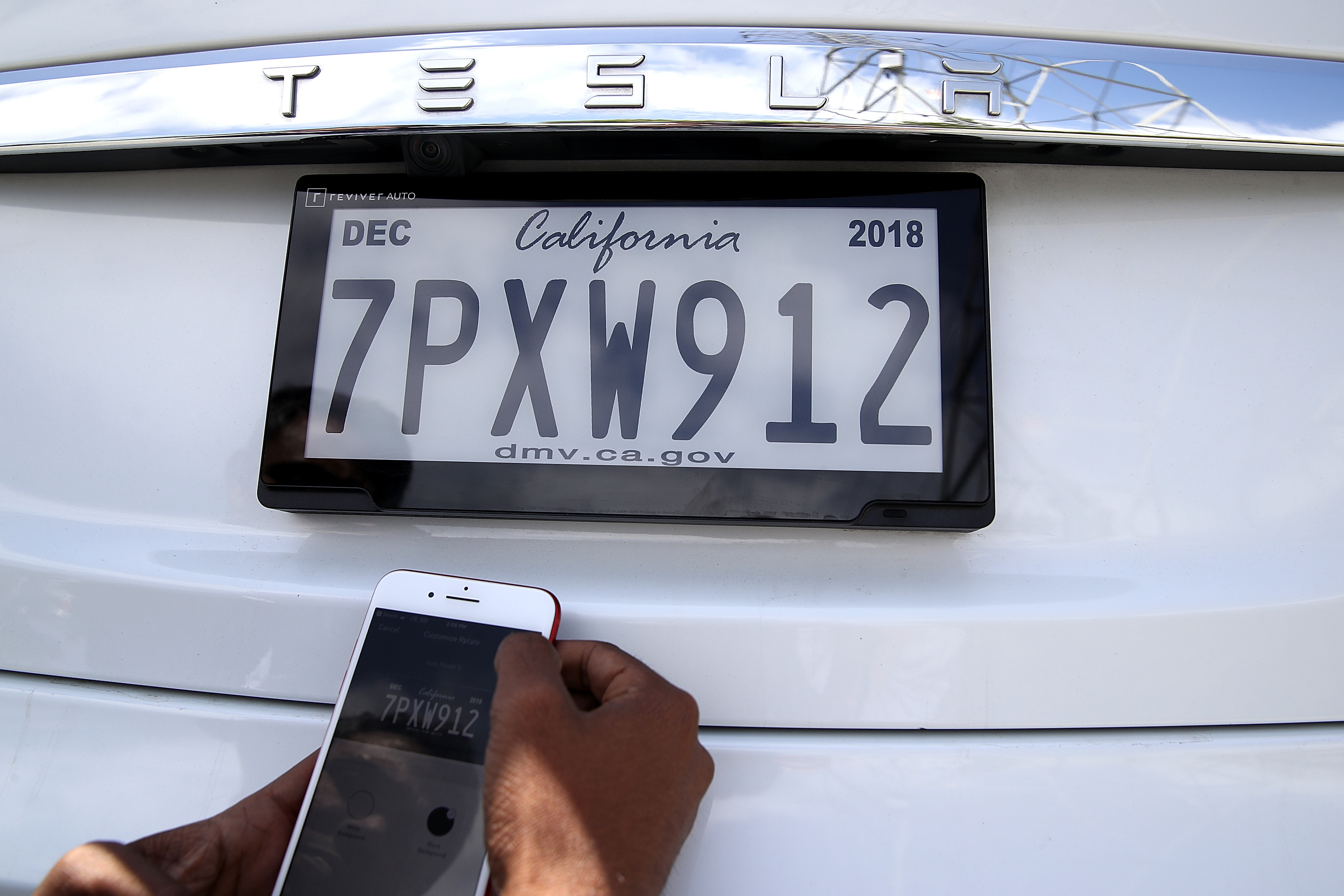 A person is holding a smartphone, matching a digital car plate on a white Tesla with "TESLA" logo above.
