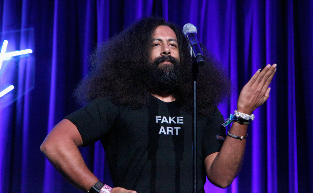 A man with a voluminous beard in a "FAKE ART" T-shirt speaks into a mic on a stage with blue lighting.