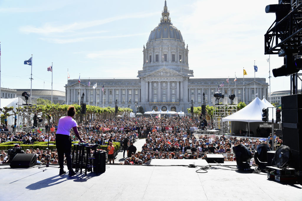 An outdoor event with a crowd facing a stage near a grand building with a dome.