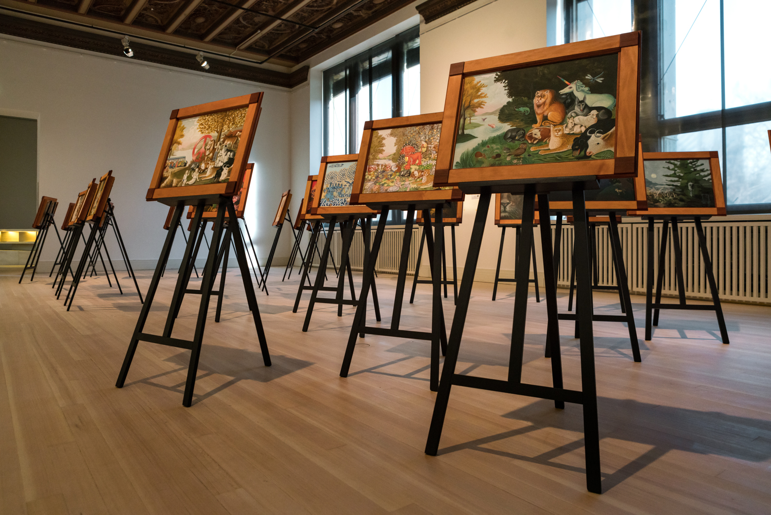 Paintings on easels in a gallery with wood floors and large windows; art appears colorful and varied.