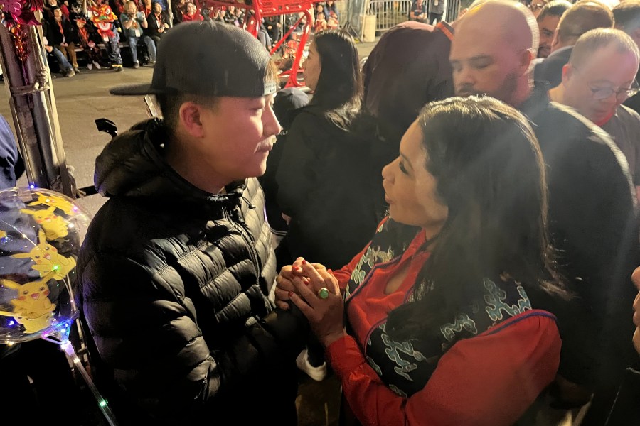 a man in a backwards black hat and a woman in a red and black jacket chat while holding hands in a crowd.